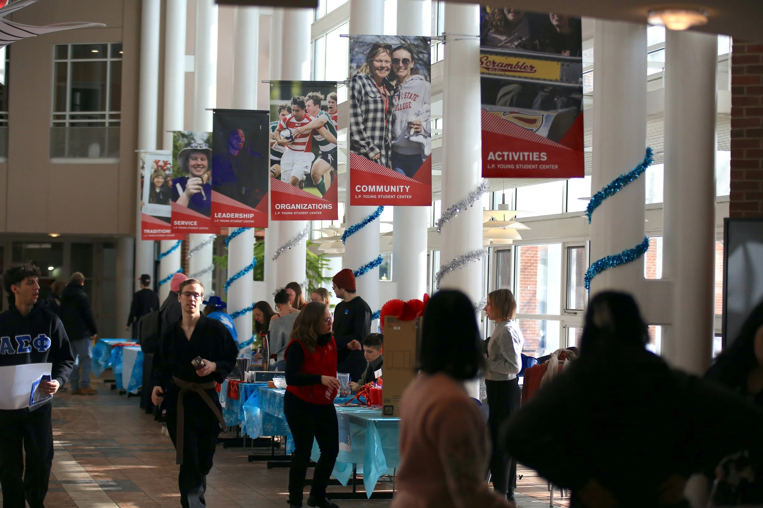 A busy indoor event with tables, people, and banners hanging from the ceiling. The banners display photos and words like 'Service', 'Leadership', 'Organizations', 'Community', and 'Activities', indicating a student center or school event. People are engaging at the tables and walking around, with some in casual or student attire.