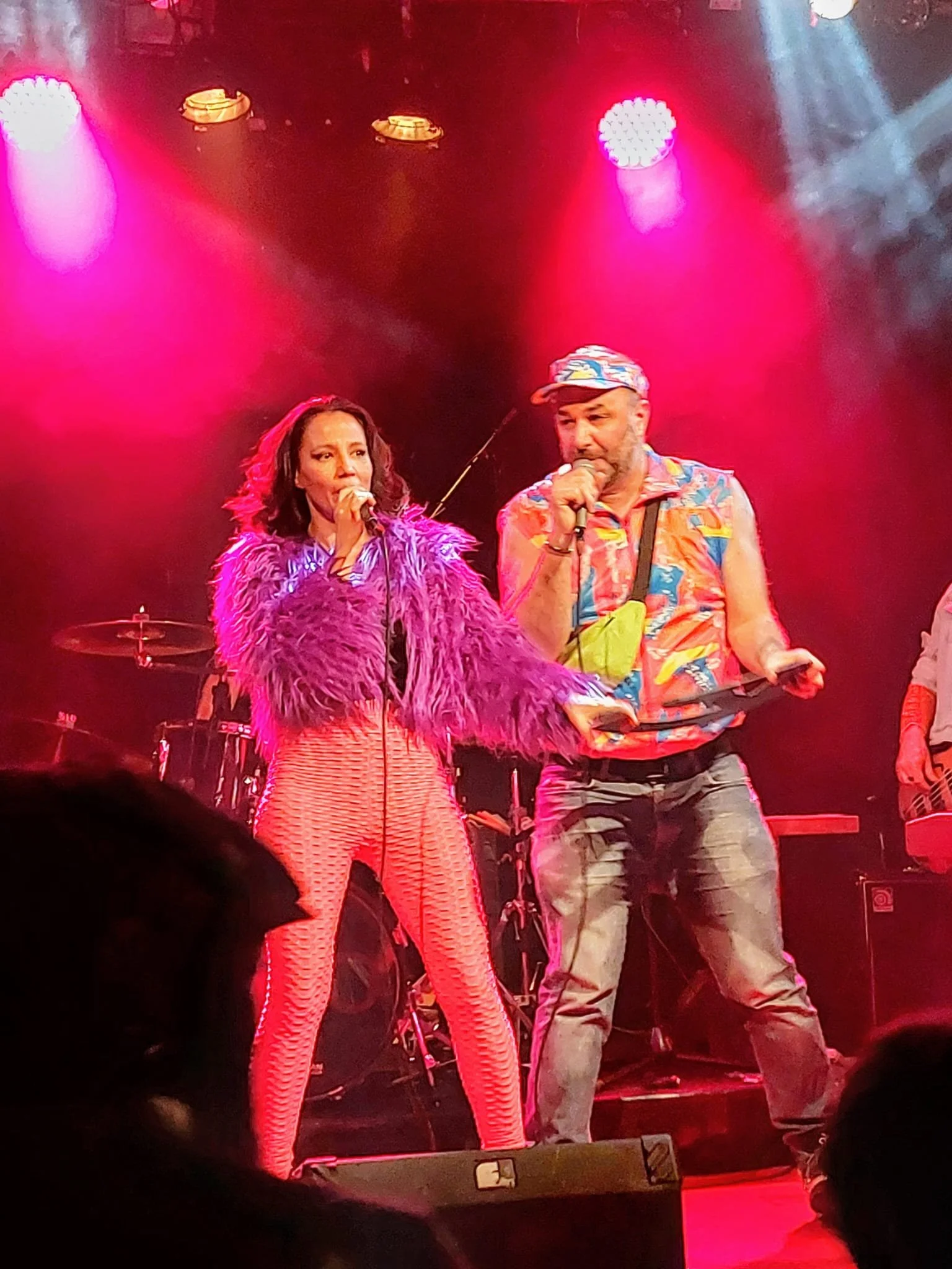Zwei Sänger auf einer Bühne bei einem Konzert, beleuchtet von pinken und weißen Scheinwerfern, mittleren Alters, ein Mann trägt ein buntes Hemd und eine Mütze, eine Frau trägt ein pinkes, pelzartiges Oberteil und passende Hosen.