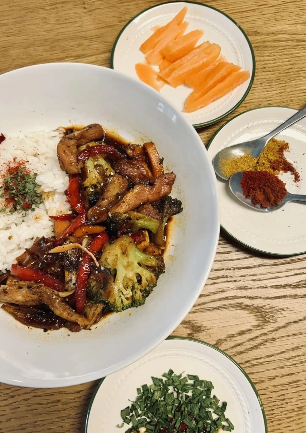 Assiette de riz blanc avec un sauté de légumes et viande, accompagnée de plusieurs petites assiettes avec des épices, du saumon fumé, et des herbes fraîches, posées sur une table en bois.
