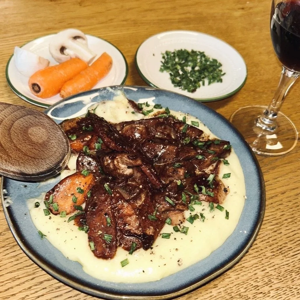 Un plat de viande avec des pommes de terre en purée, accompagné de légumes (carottes et champignons) et d'un verre de vin rouge.