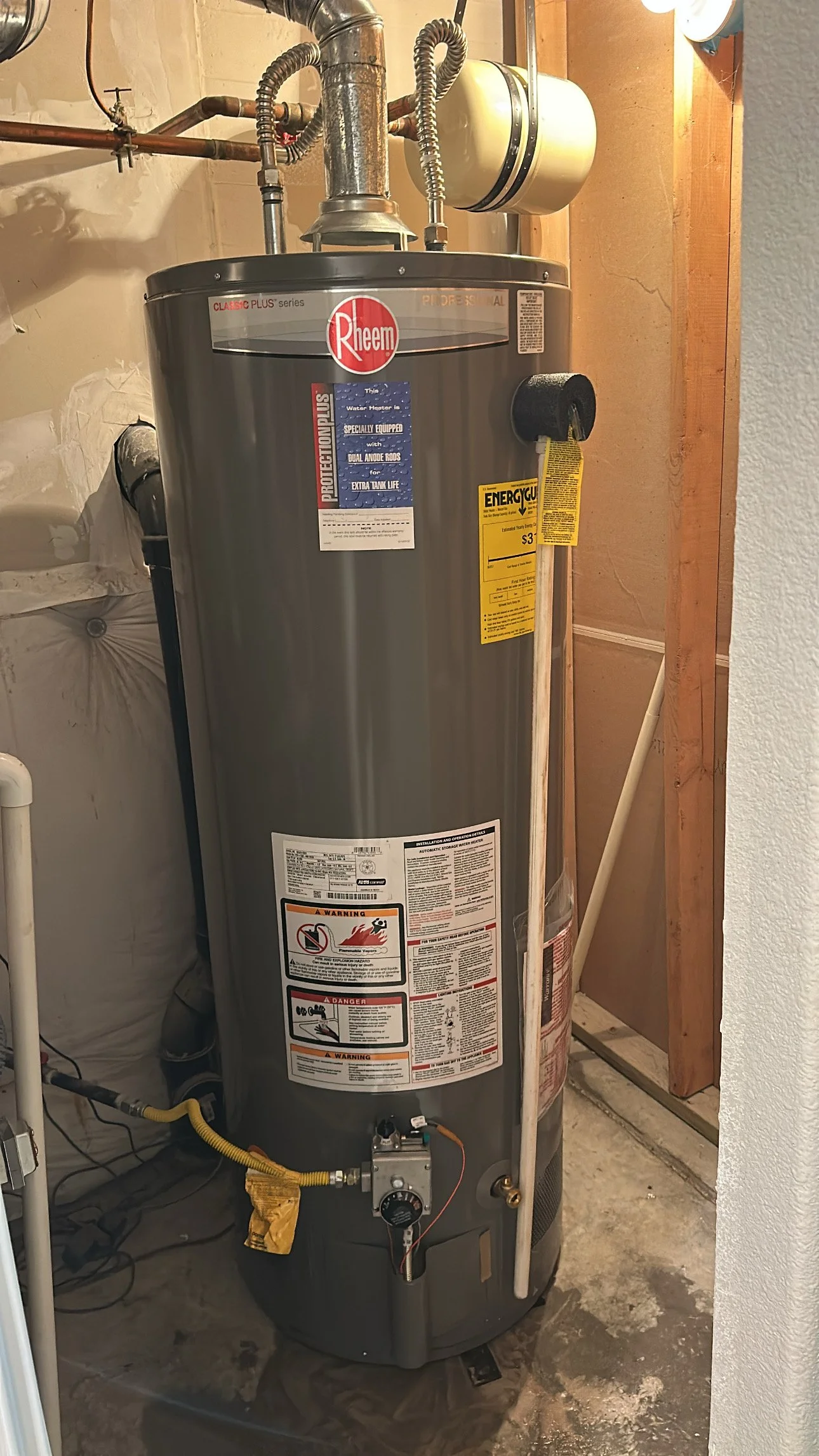 A tall, gray Rheem water heater situated in a utility room with exposed plumbing and electrical connections.