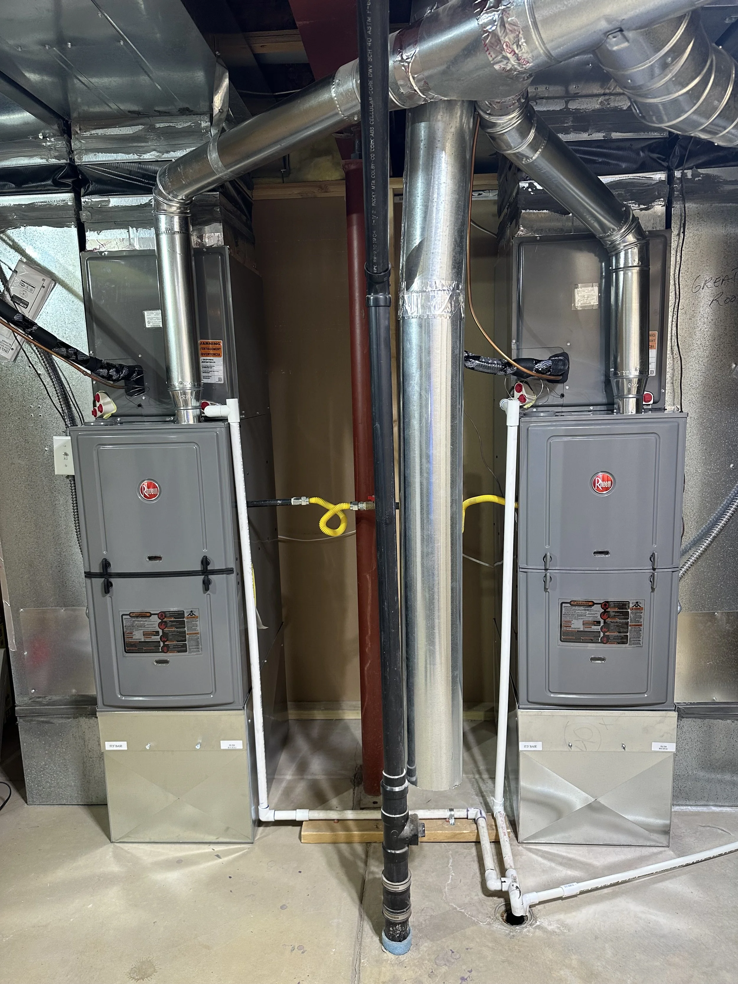 Two HVAC furnaces with metal ducts, pipes, and venting system in a basement or utility room.