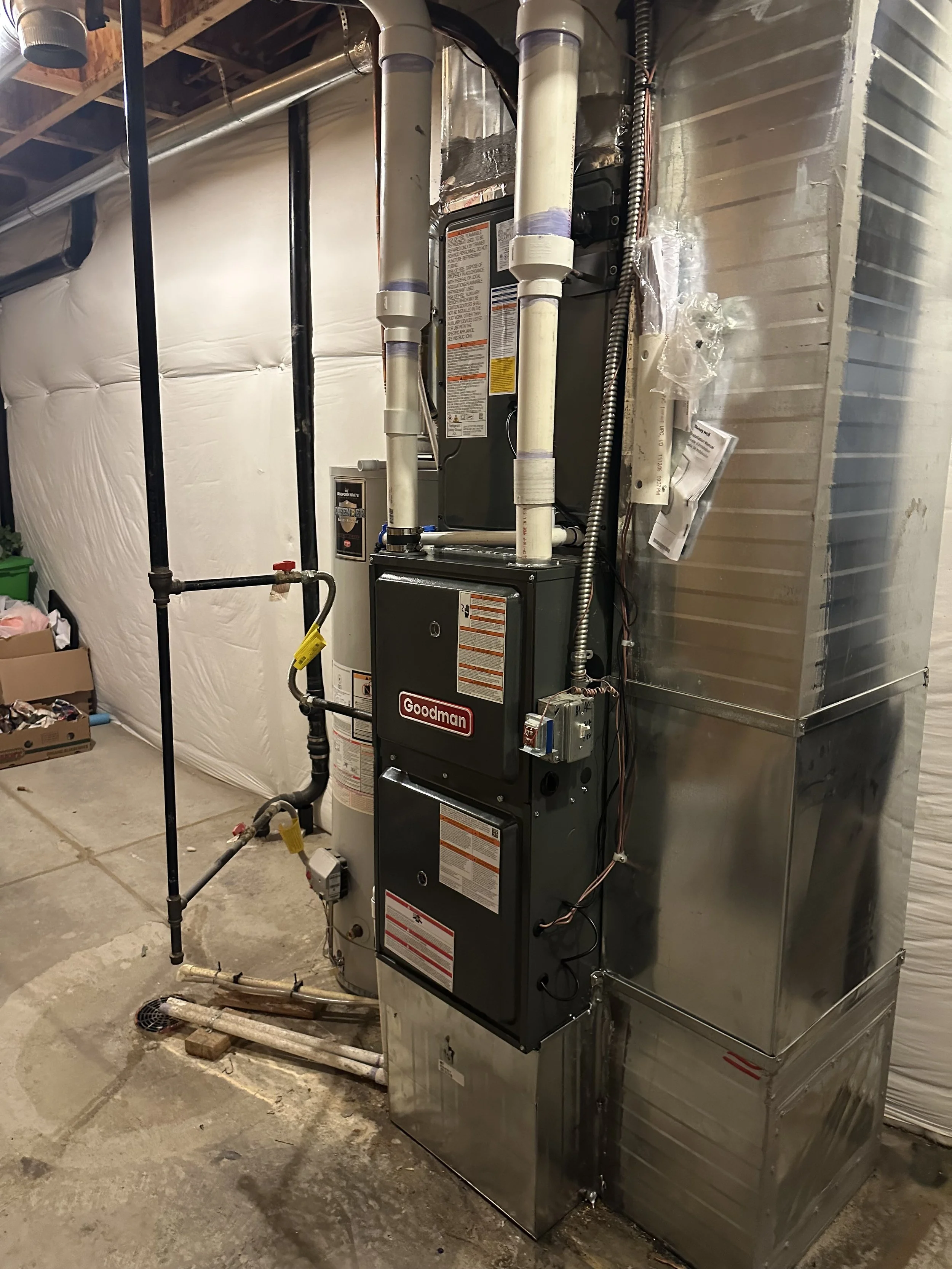 A Goodman furnace and duct system installed in a basement, with PVC pipes, electrical wiring, and a concrete floor.