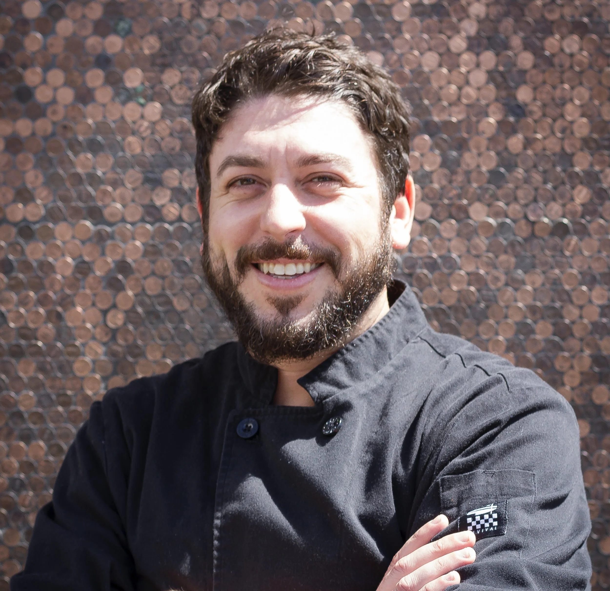 A smiling man with a beard and dark hair, wearing a black chef's coat, standing in front of a textured copper wall.