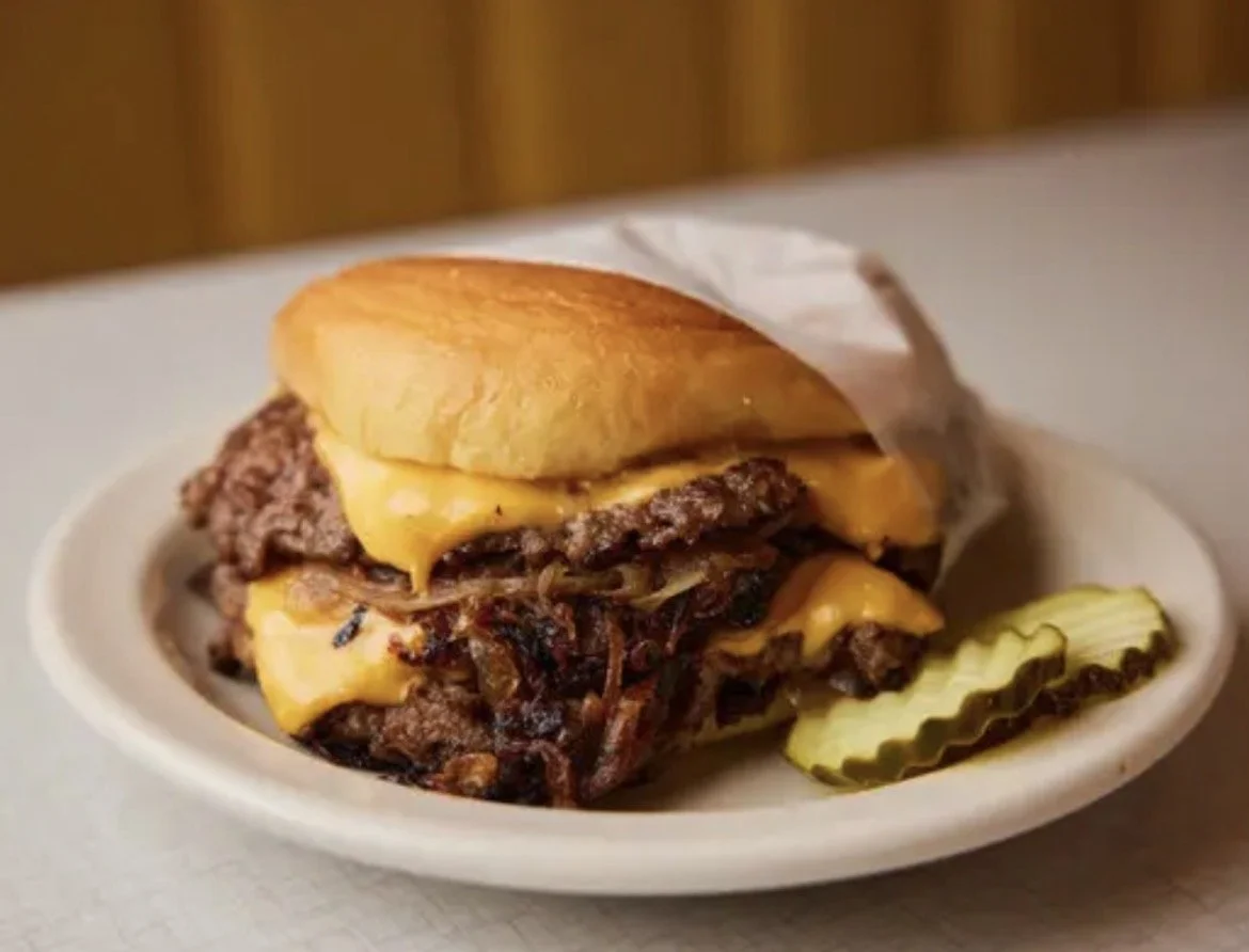 A cheeseburger with two beef patties, melted cheddar cheese, and a toasted bun, served with pickles on the side.