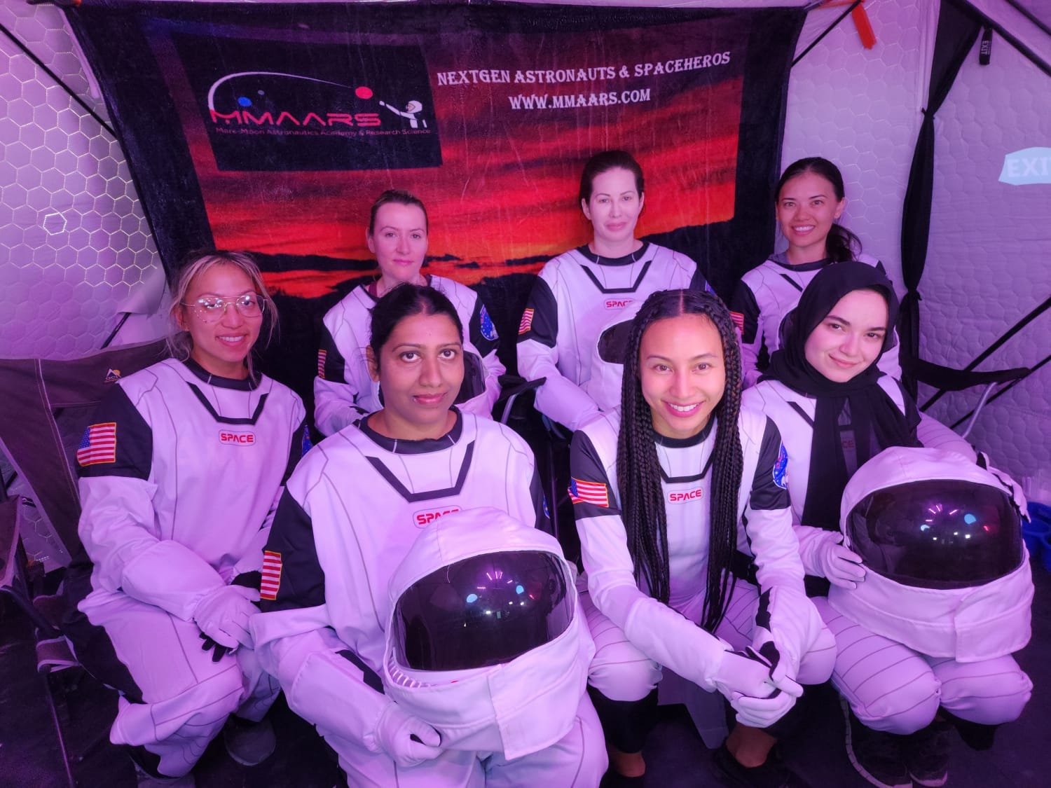 Group of eight women dressed as astronauts in space suits, sitting and posing for a photo in front of a space-themed backdrop with the words "Nextgen Astronauts & Spaceheroes" and a website URL.