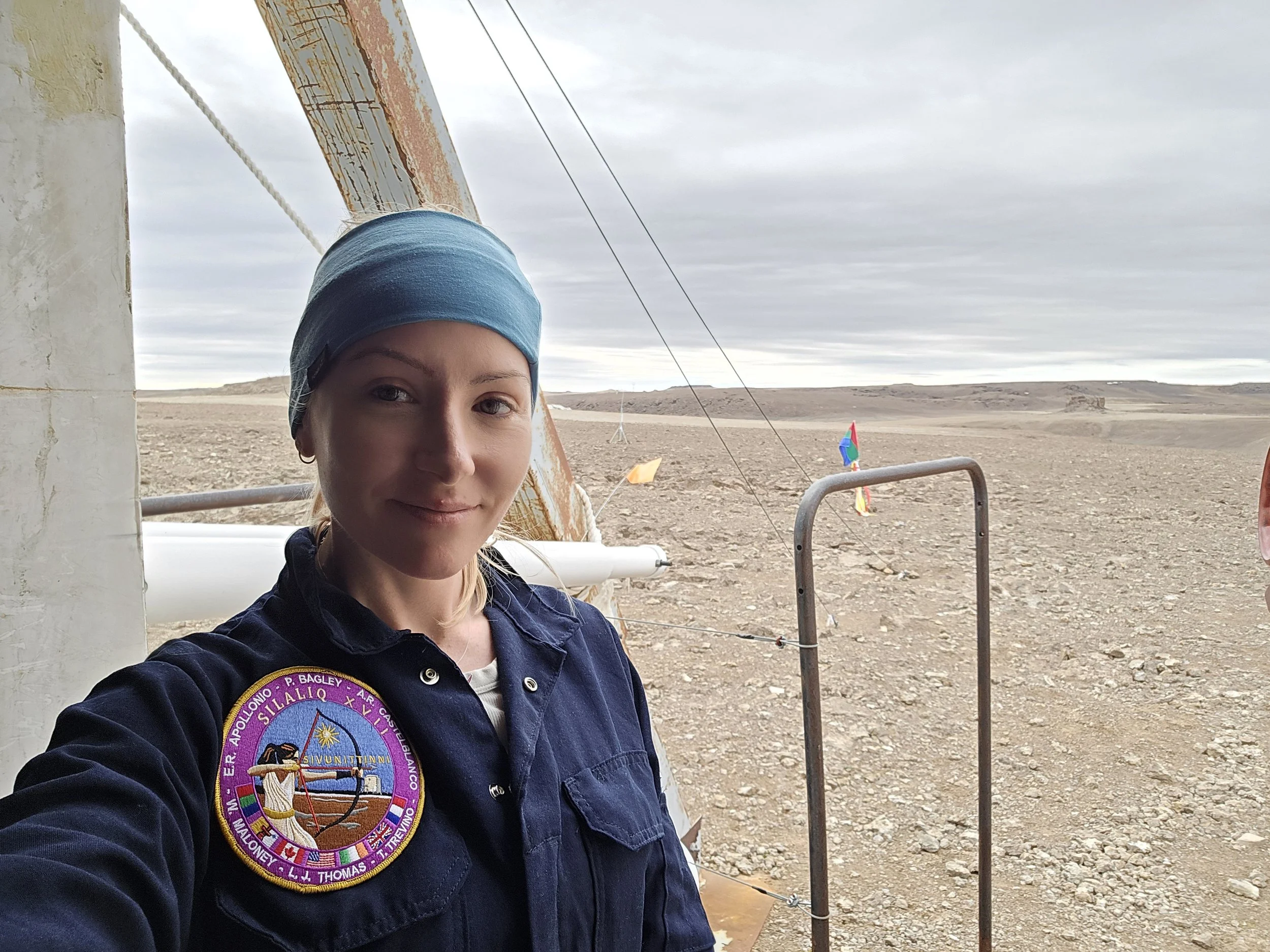A woman in a navy uniform with a NASA patch on her shoulder takes a selfie in a desert-like environment with a cloudy sky. In the background, there are sparse plants, flags, and a metal rail fence.