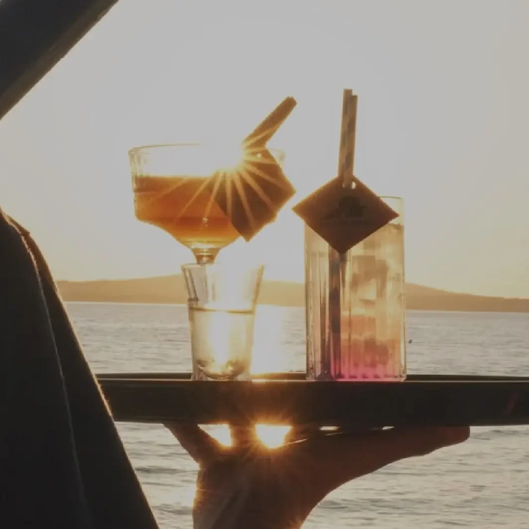 Two vibrant, colorful cocktails on a tray with a blurry Zadar sunset and beach club background.