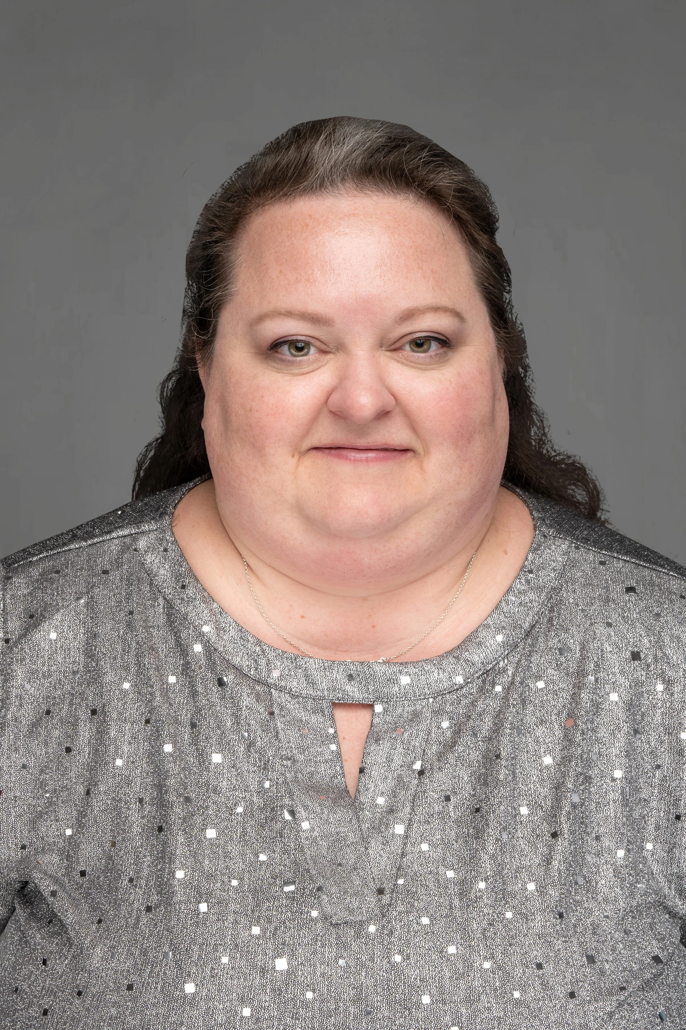 Portrait of a woman with long dark hair wearing a sparkly silver top with small black and white squares, against a gray background.