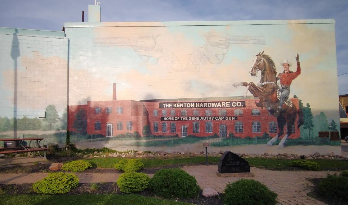 Mural on building depicting a cowboy riding a horse, with a brick factory in the background, and text that reads 'The Kenton Hardware Co. Home of the Gene Autry Cap Gun'.