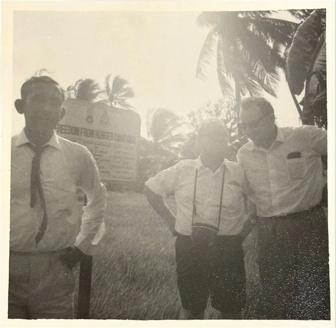 Three men standing outdoors, with palms and a billboard in the background, in black and white.