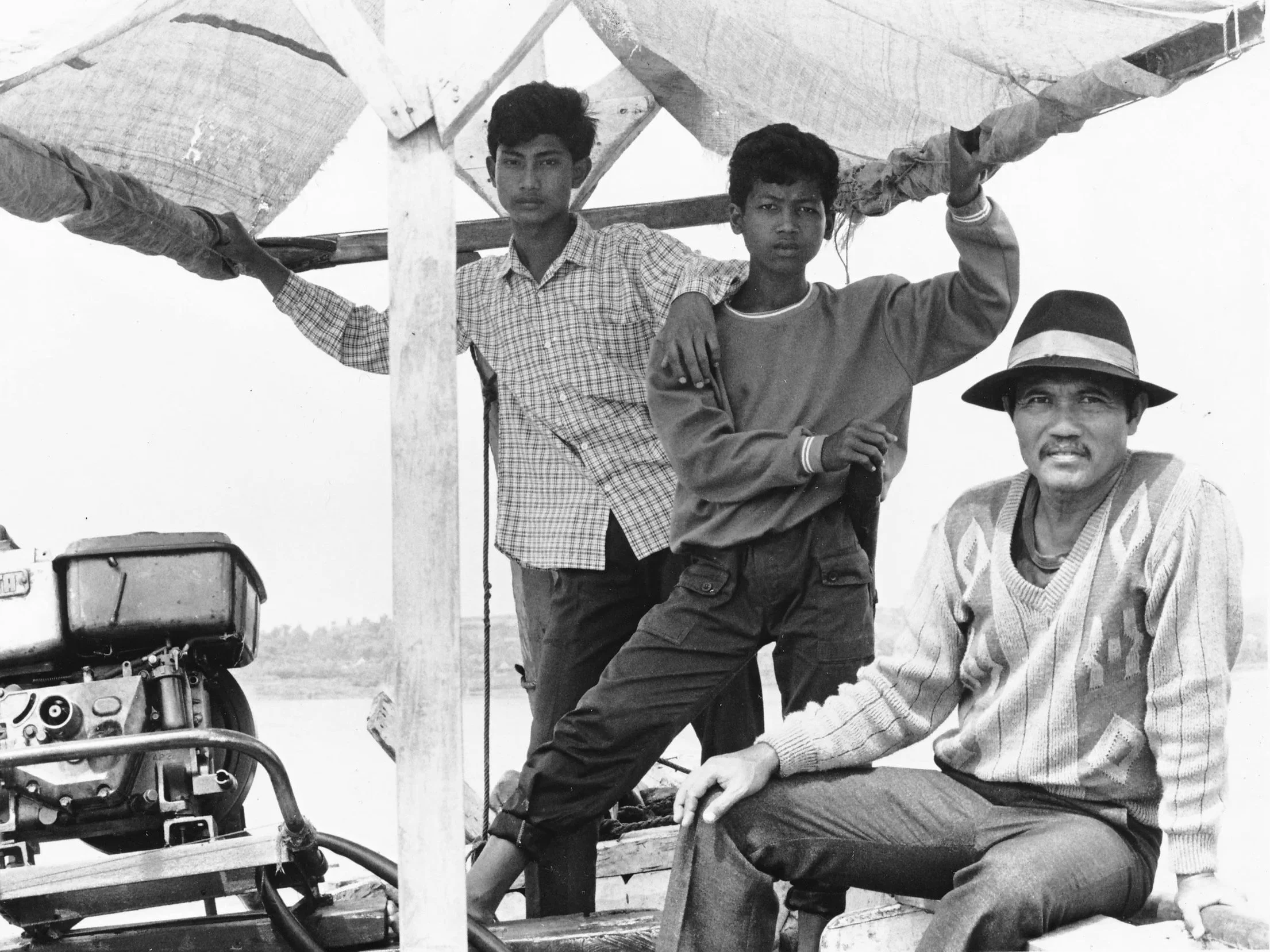 Black and white photo of two boys and a man on a boat. One boy is wearing a checked shirt. The man is wearing a hat. The background is open sky.