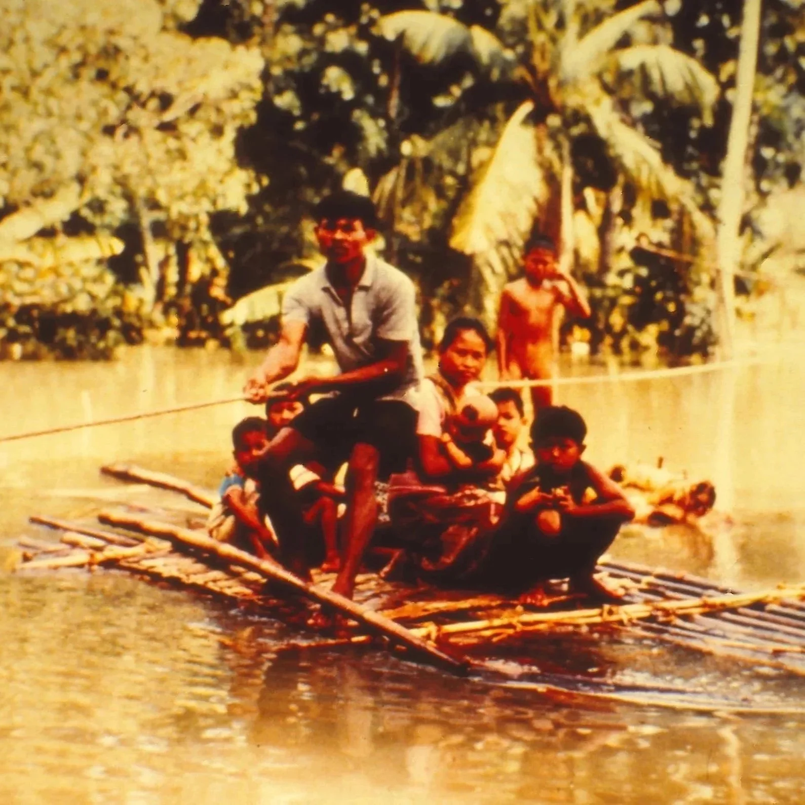 A group of people on a bamboo raft floating on a river in a tropical setting with banana trees in the background.