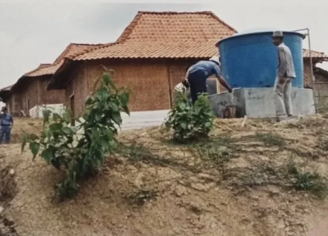 A men working near a large blue water tank in front of wooden buildings with red-tiled roofs, on a dirt hill with small green plants.