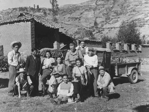A group of people posing outdoors near a brick building and a truck, with hills in the background.