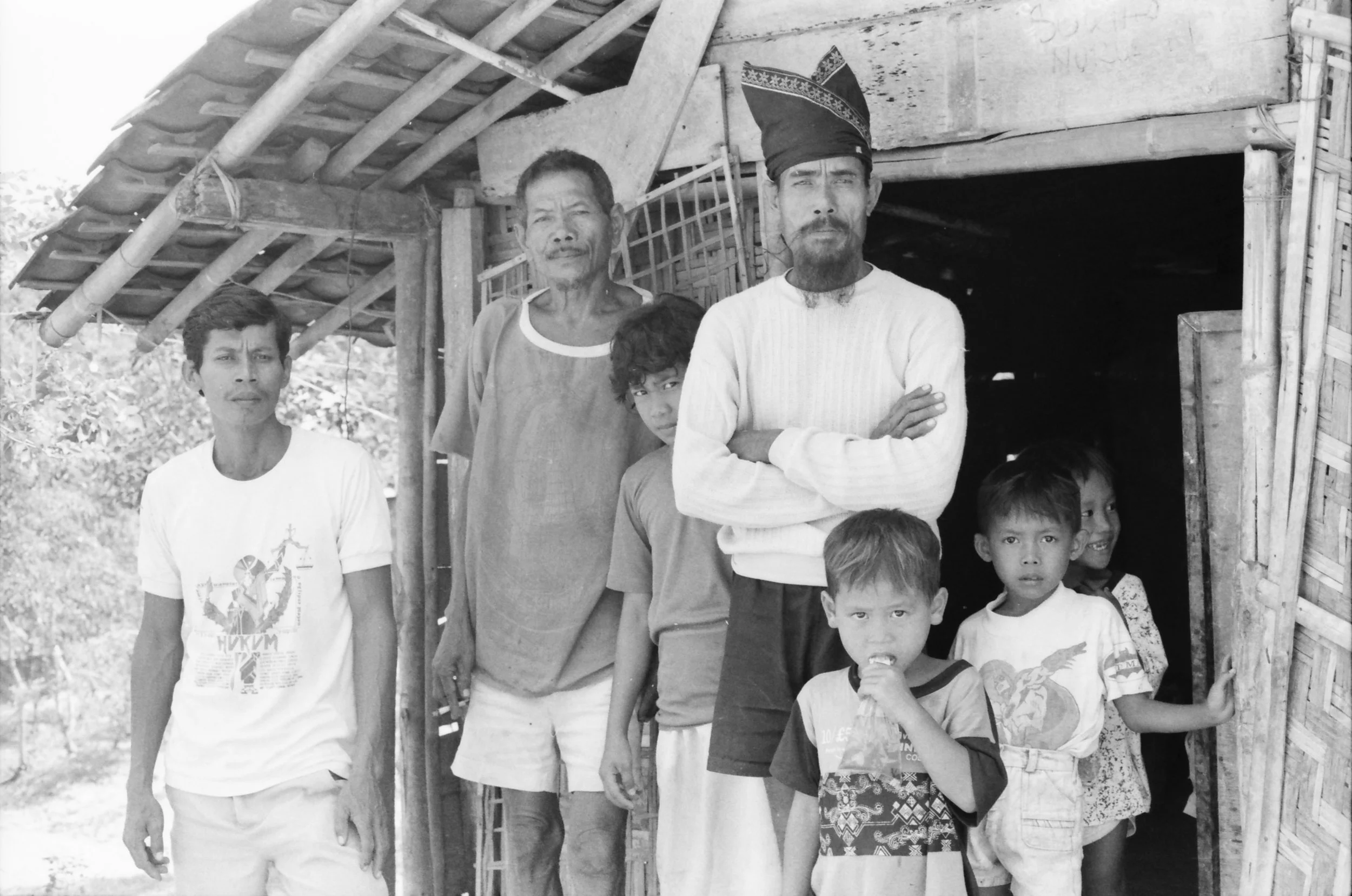 A group of men and children standing in front of a wooden structure. The men are of varying ages, some with facial hair and distinctive clothing, one wearing a traditional hat. Children are standing at the front, some smiling.
