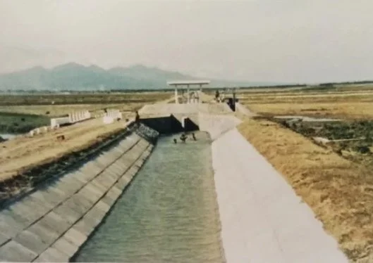 View of a dam or water control structure with a concrete spillway and a gate station, surrounded by flat agricultural land and distant mountains.