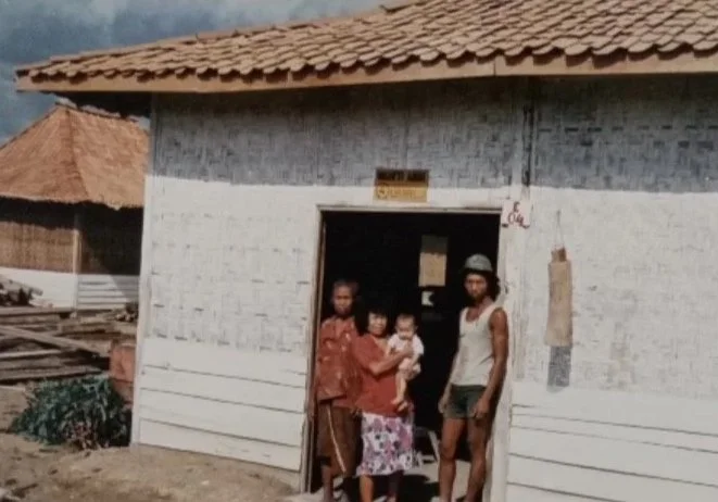 Four people standing outside a rustic house. One woman is holding a child, and there are two men standing nearby.