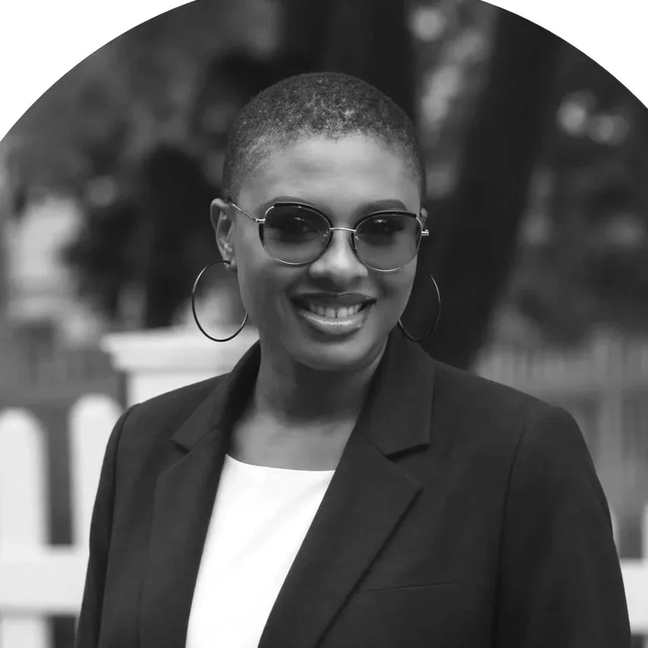 A photo of Chiamaka Uzomba, smiling with a short haircut wearing sunglasses, hoop earrings, a dark blazer, and a white shirt, outdoors with trees and a fence in the background.