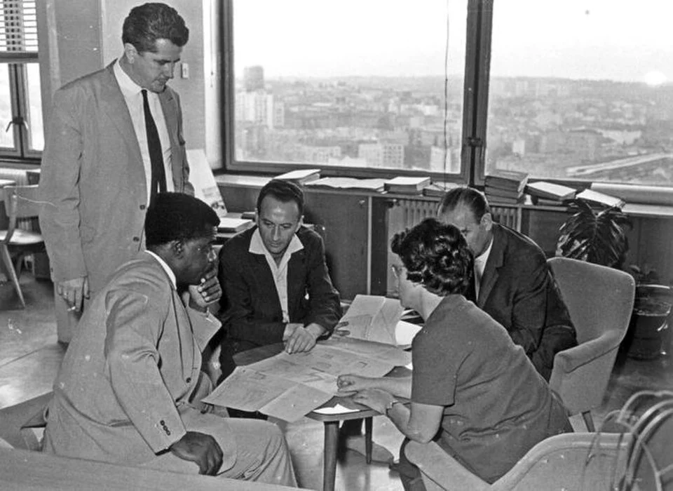 Five people are gathered around a table in an office, reviewing documents. One man is standing, while the others sit and look at papers, with large windows showing a cityscape in the background.