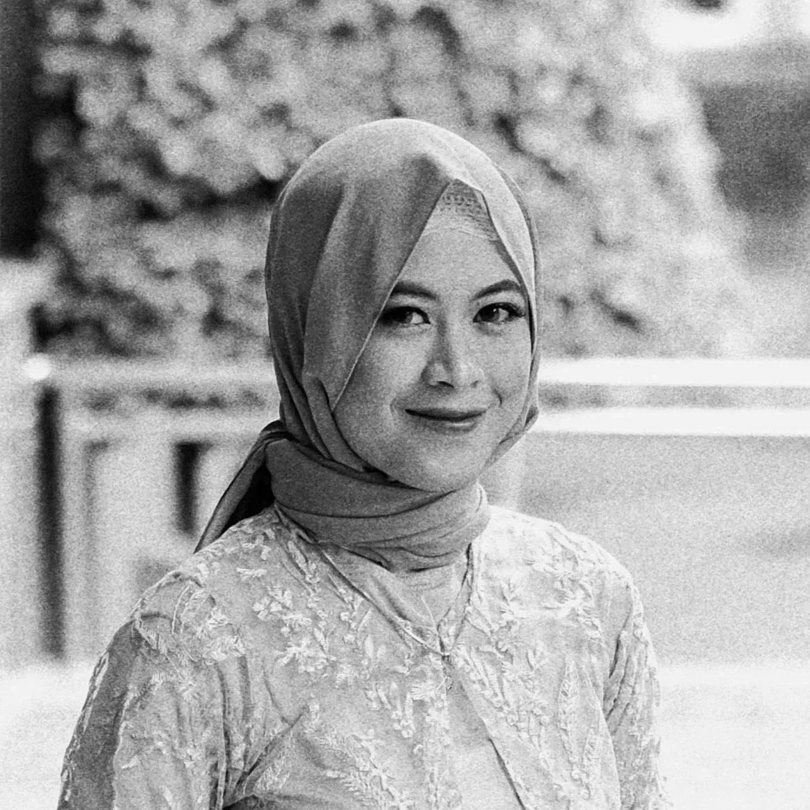 Black and white photo of Syarifah Aini, wearing a headscarf and traditional embroidered dress, smiling outdoors.