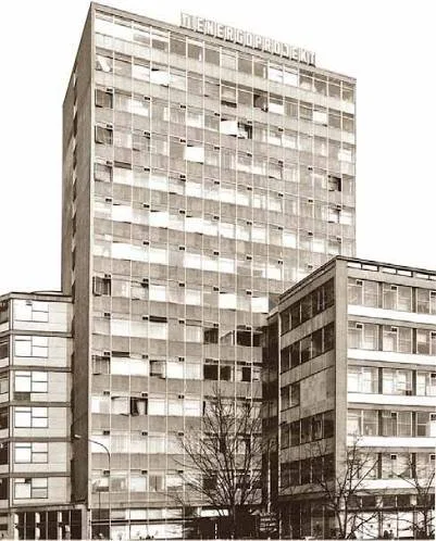 A black and white photo of a large multi-story office building with a sign in Russian on top. The building has many windows, some open, and is adjacent to a smaller building. There are trees and streetlights in front.