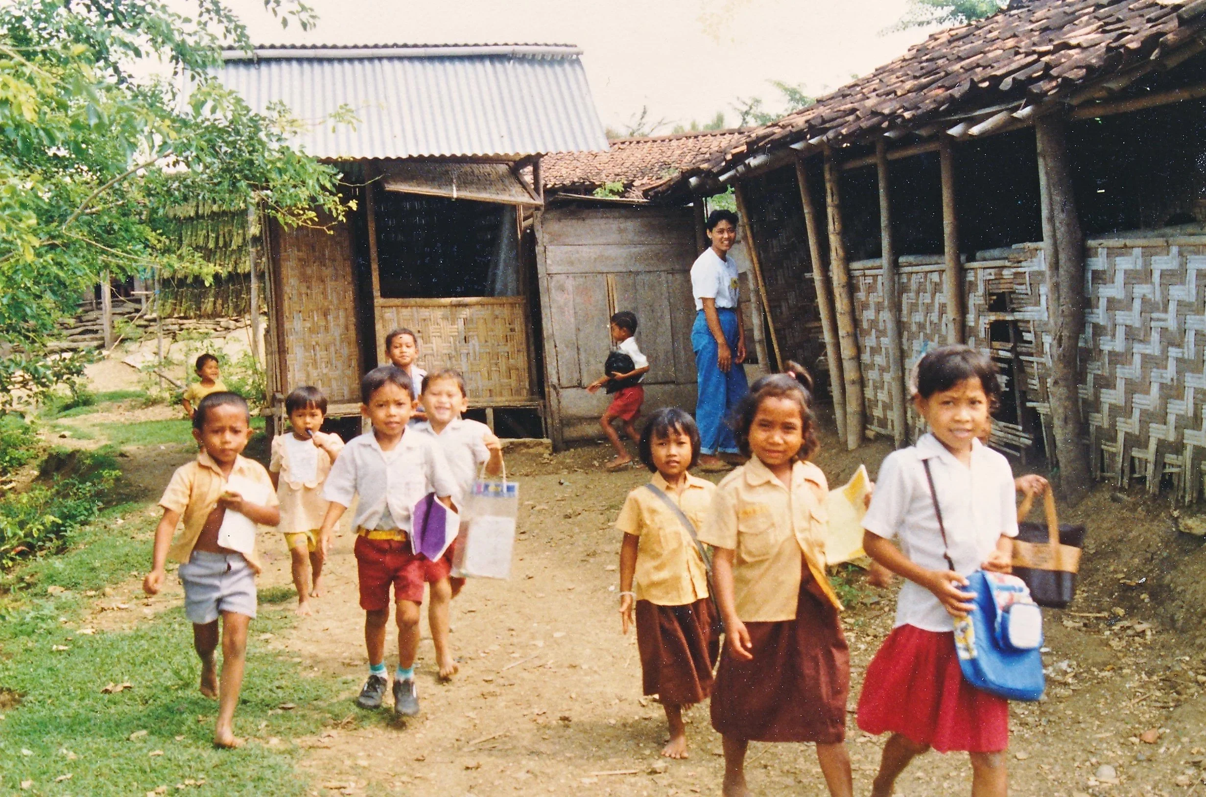 A group of children walking outside near traditional wooden houses in a rural area. The children are smiling and carrying books or bags. The background shows some greenery and trees.