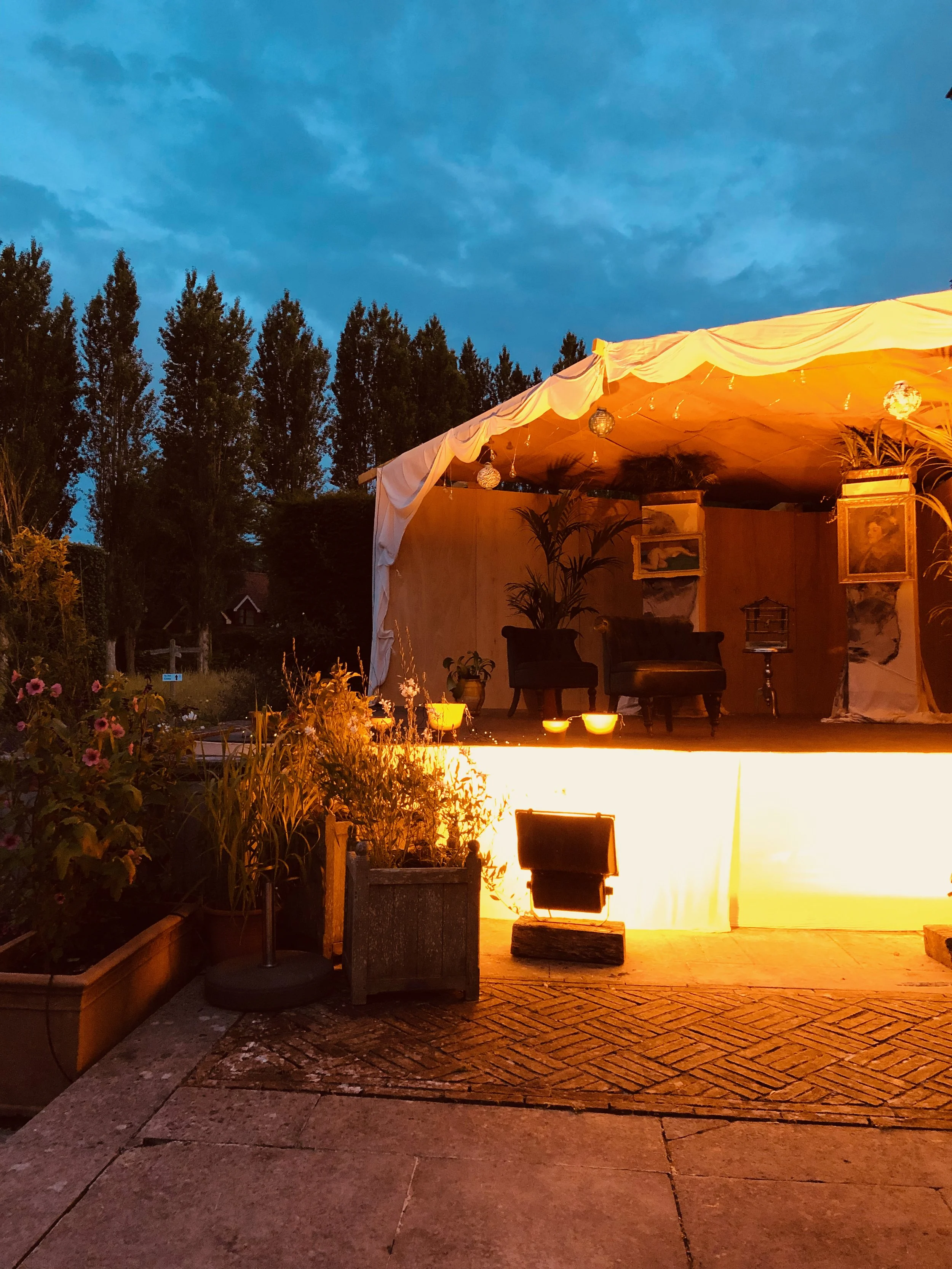 An outdoor stage with warm lighting, decorated with plants and hanging lights, set against a background of trees and a twilight sky.