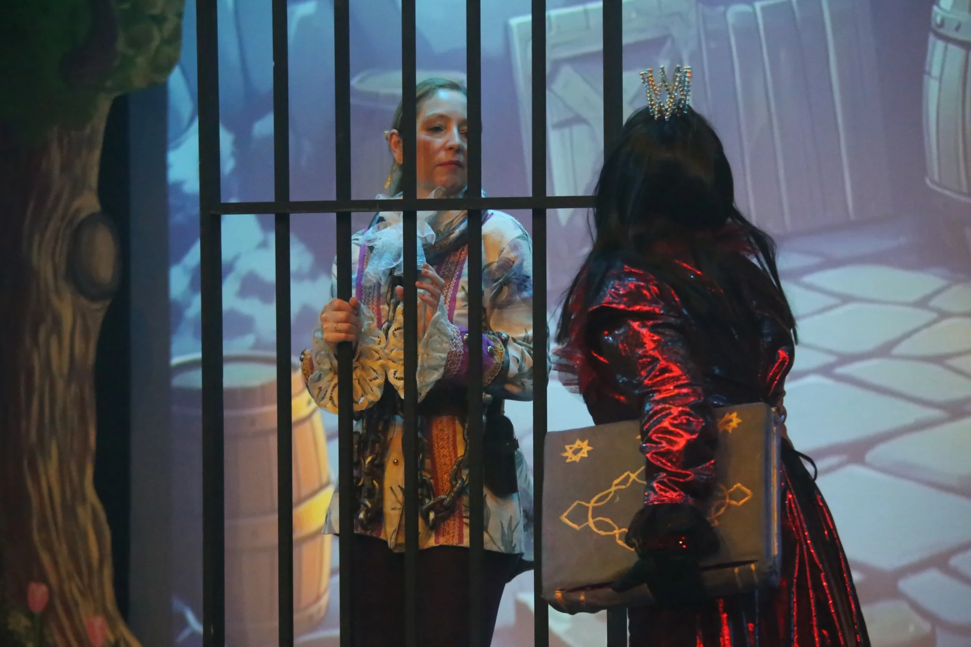 Two women on stage, one behind bars, the other facing away holding a box, with colorful background imagery.