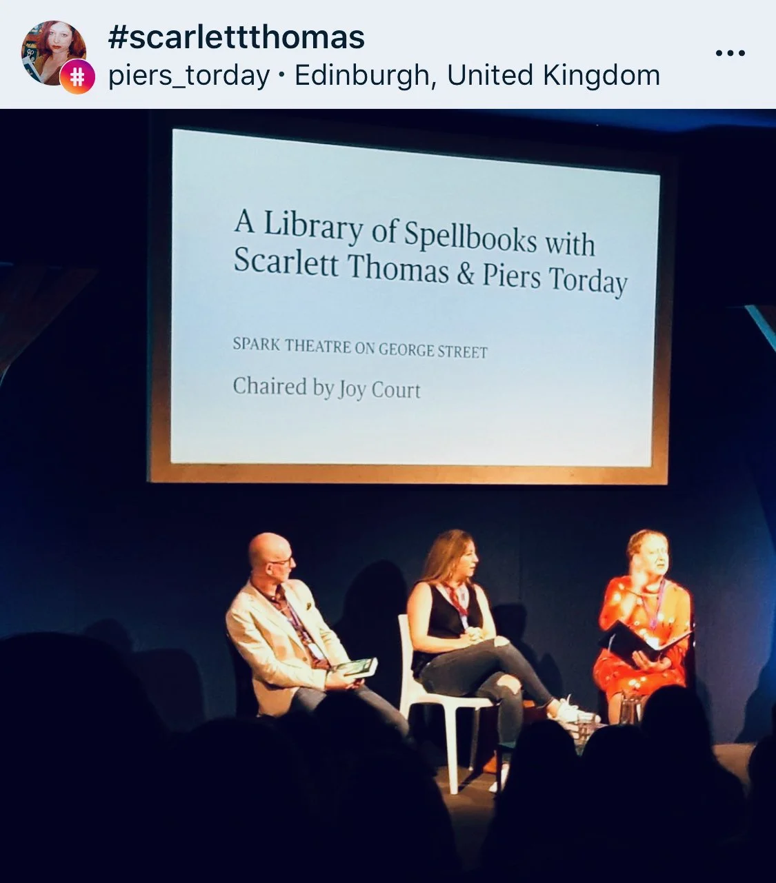 Panel discussion at a theatre with three speakers, a large screen displaying information about a library of spellbooks with Scarlett Thomas and Piers Torday. Audience silhouette in foreground.