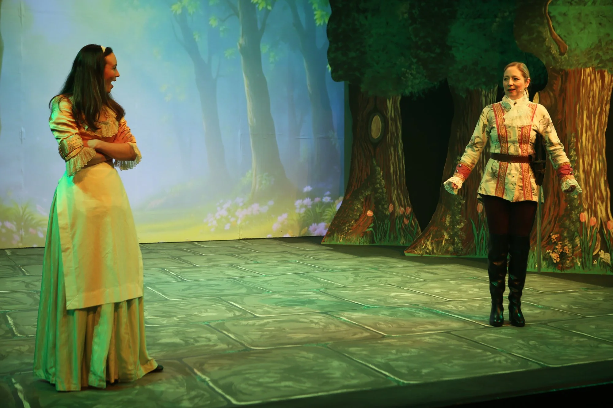 Two women in period costumes standing on a stage with a forest scene background. One woman is smiling with her arms crossed, wearing a long yellow dress. The other woman, dressed in a patterned jacket, black pants, and tall black boots, is standing w