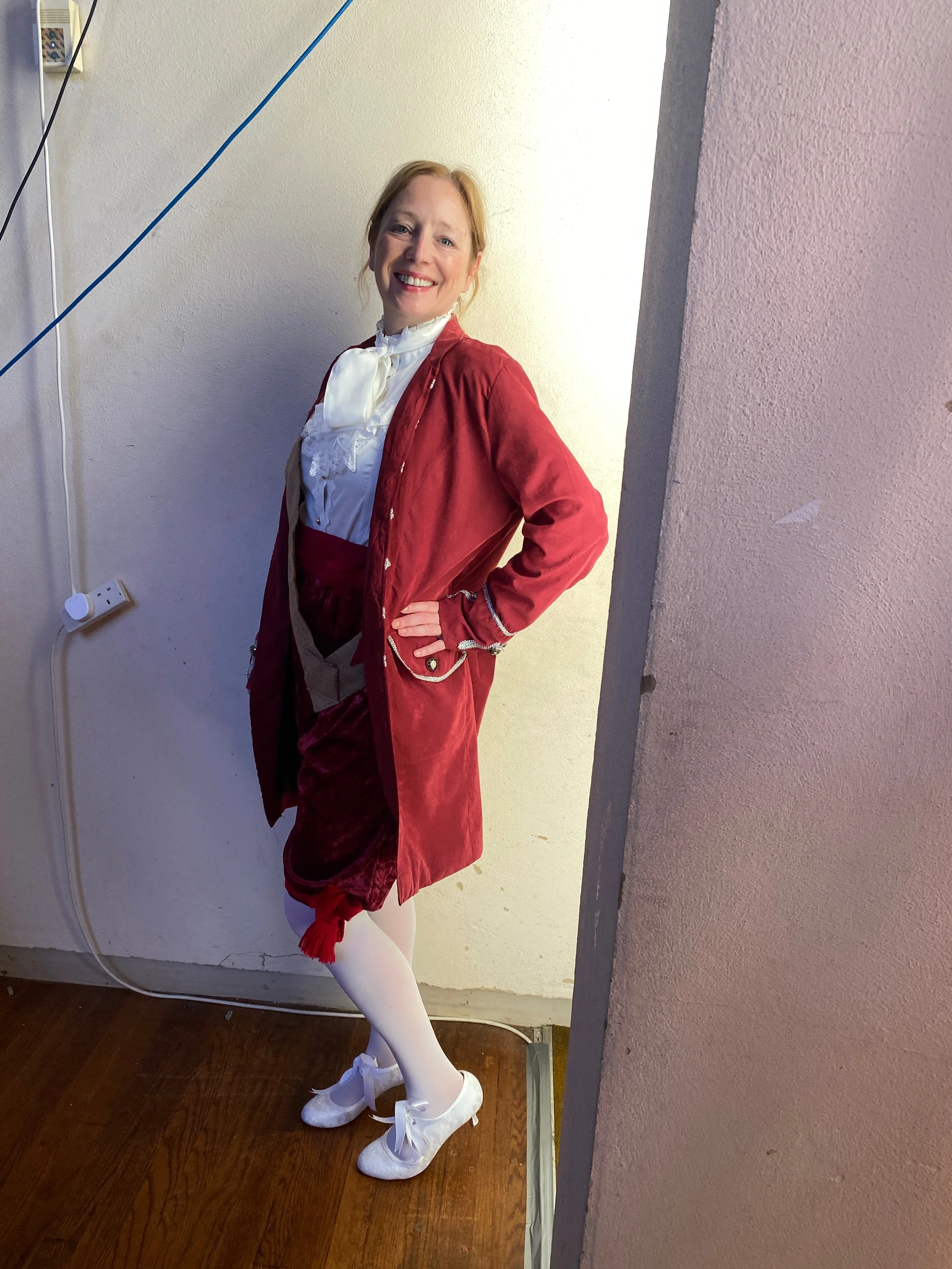 A woman dressed in historic 18th-century style clothing, including a red velvet coat, white lace shirt, burgundy skirt, white tights, and white shoes, standing indoors and smiling at the camera.