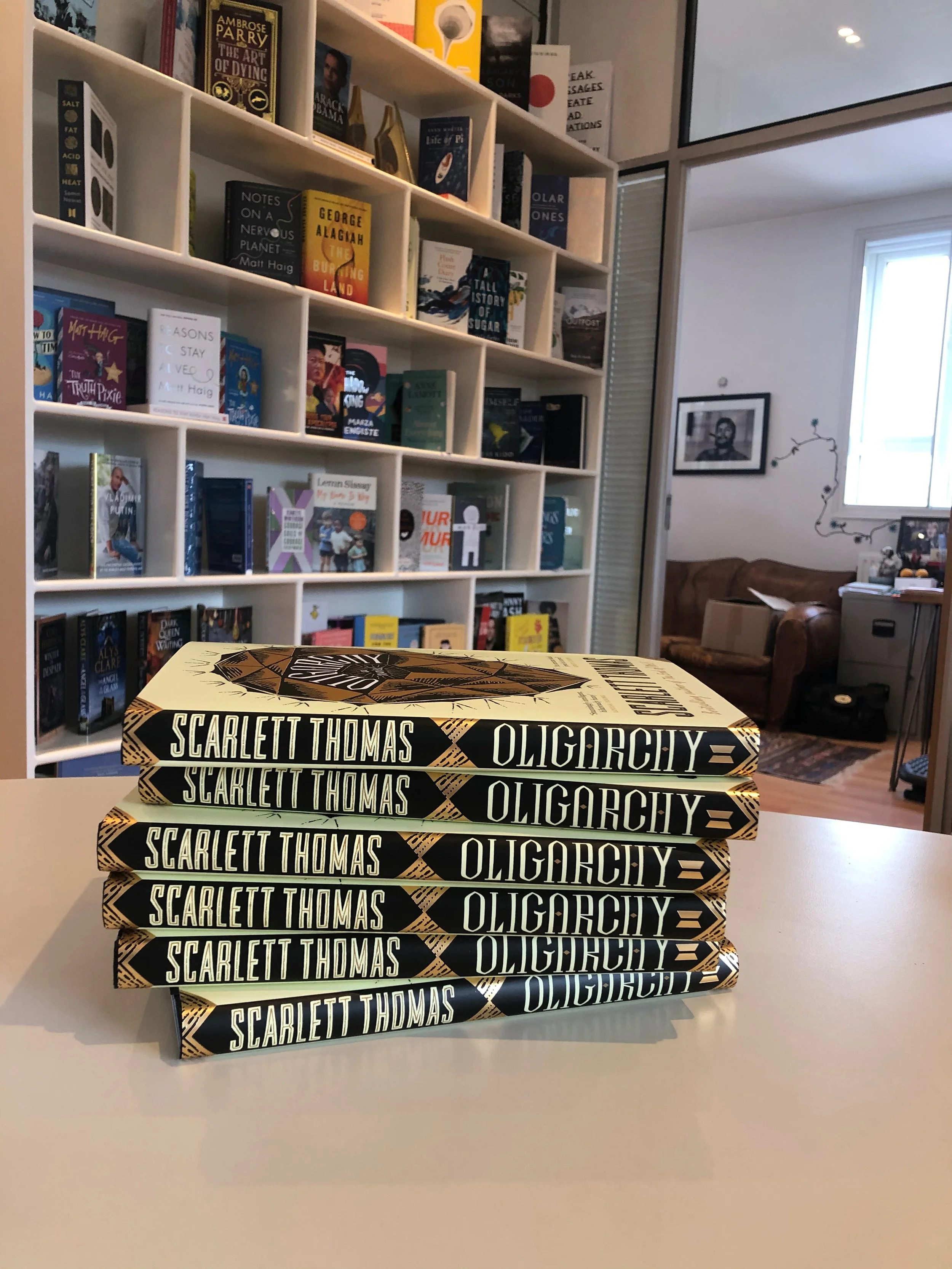 A stack of five books titled "Oligarchy" by Scarlett Thomas on a white table in a bookstore or library. In the background, white shelves filled with various books and a cozy seating area with a brown couch and a window.