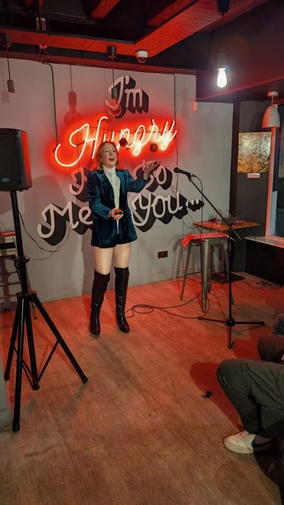 A woman singing or speaking into a microphone on a small stage in a bar or restaurant with a neon sign that says 'I'm Happy to Meet You' in the background.