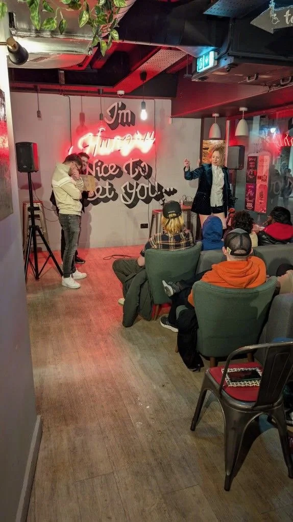 A woman is performing on stage in a cafe or bar, with a small audience seated and watching. Behind her, a wall has a neon sign that reads 'I'm Always Nice To Get You...'. The stage setup includes speakers and a microphone. The setting has a cozy, mod