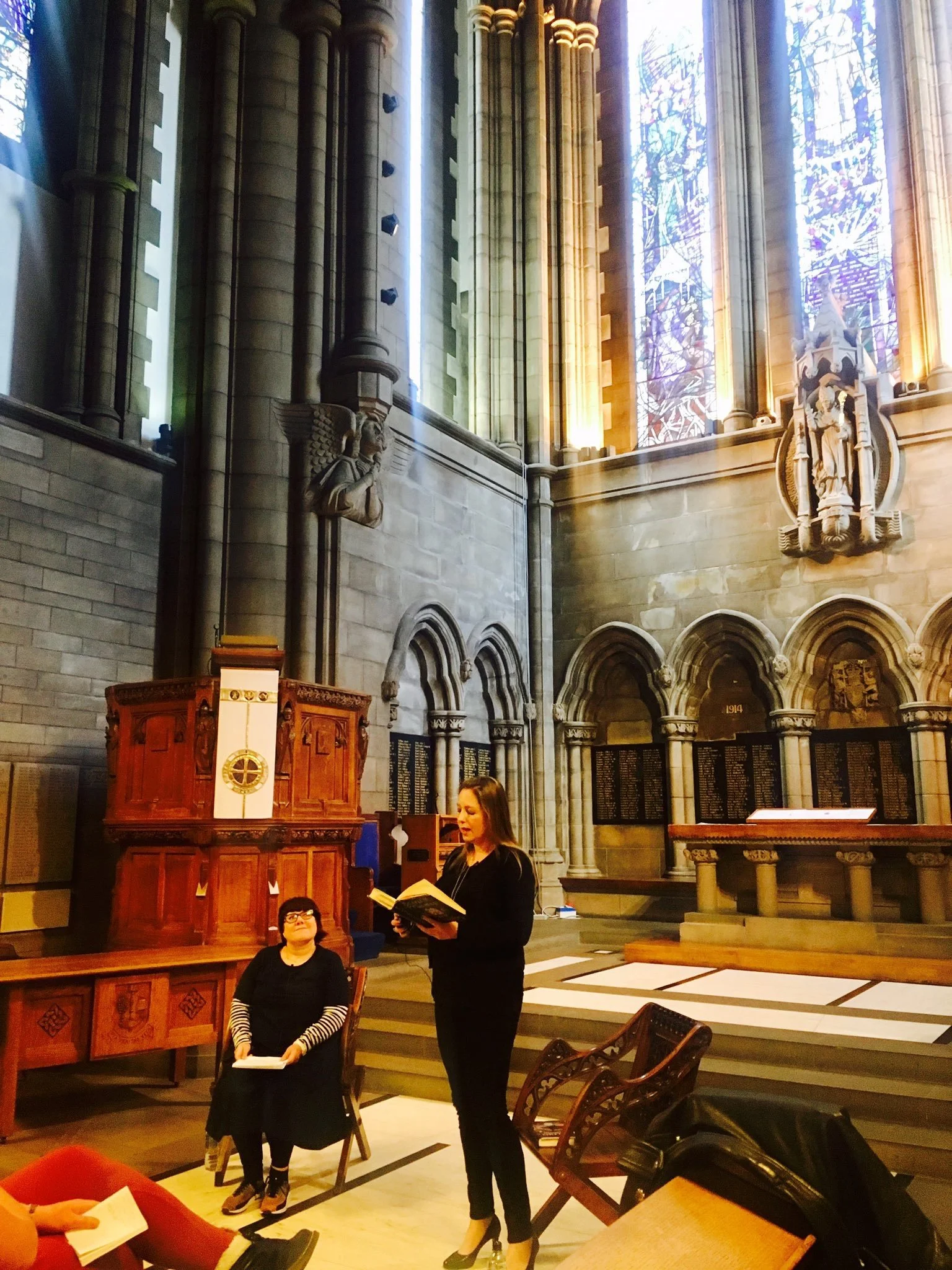 Two women are inside a large, ornate church with high stained glass windows. One woman stands reading from a book, while the other is seated looking up at her. The church features stone walls, arched windows, sculptures, and wooden furniture.