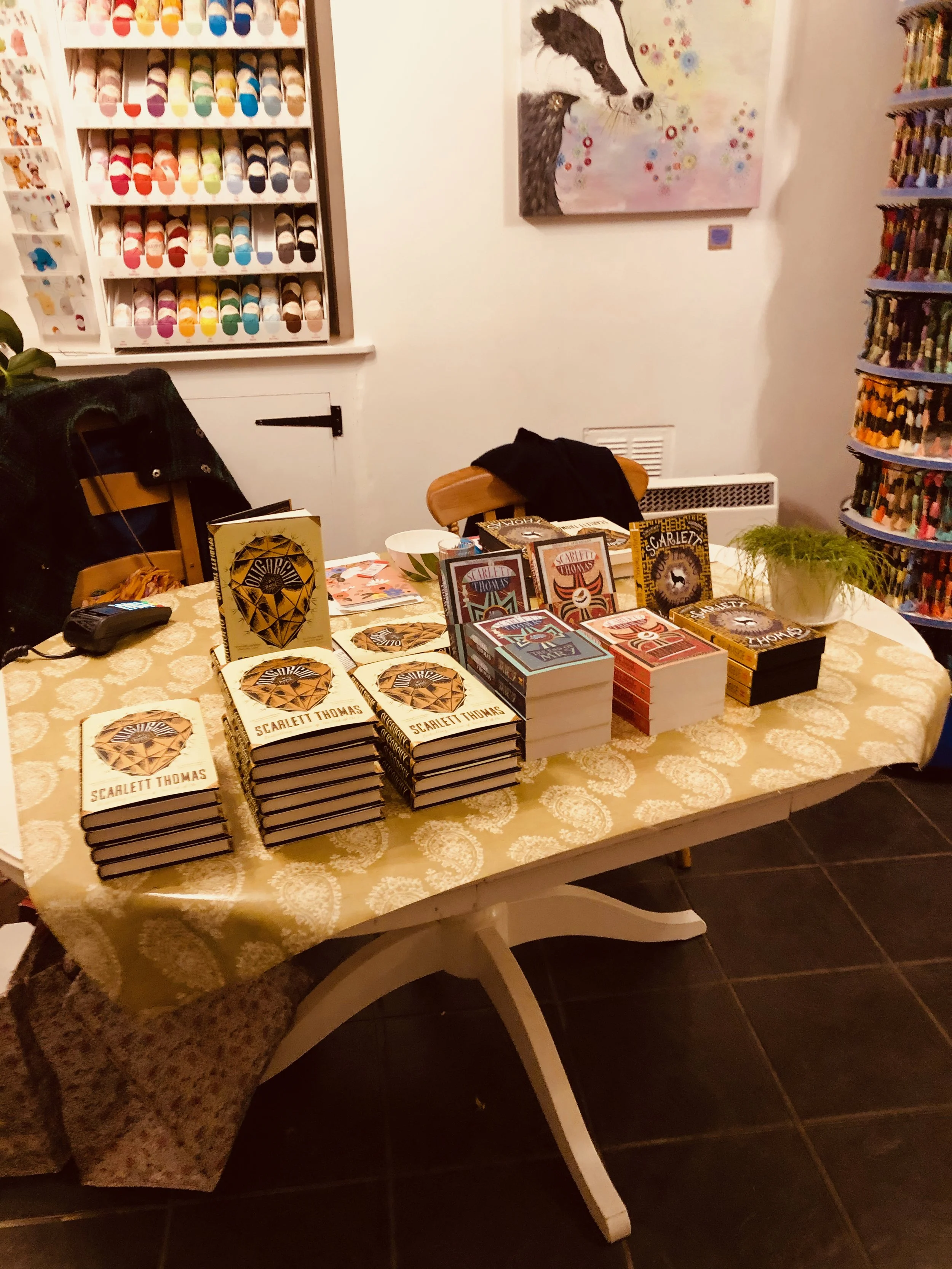 A table covered with a patterned tablecloth displays stacks of books by Scarlett Thomas. The background shows shelves of colorful yarn balls, a painting of a stylized horse, and a round display of colored pencils. There is a chair with a black item d