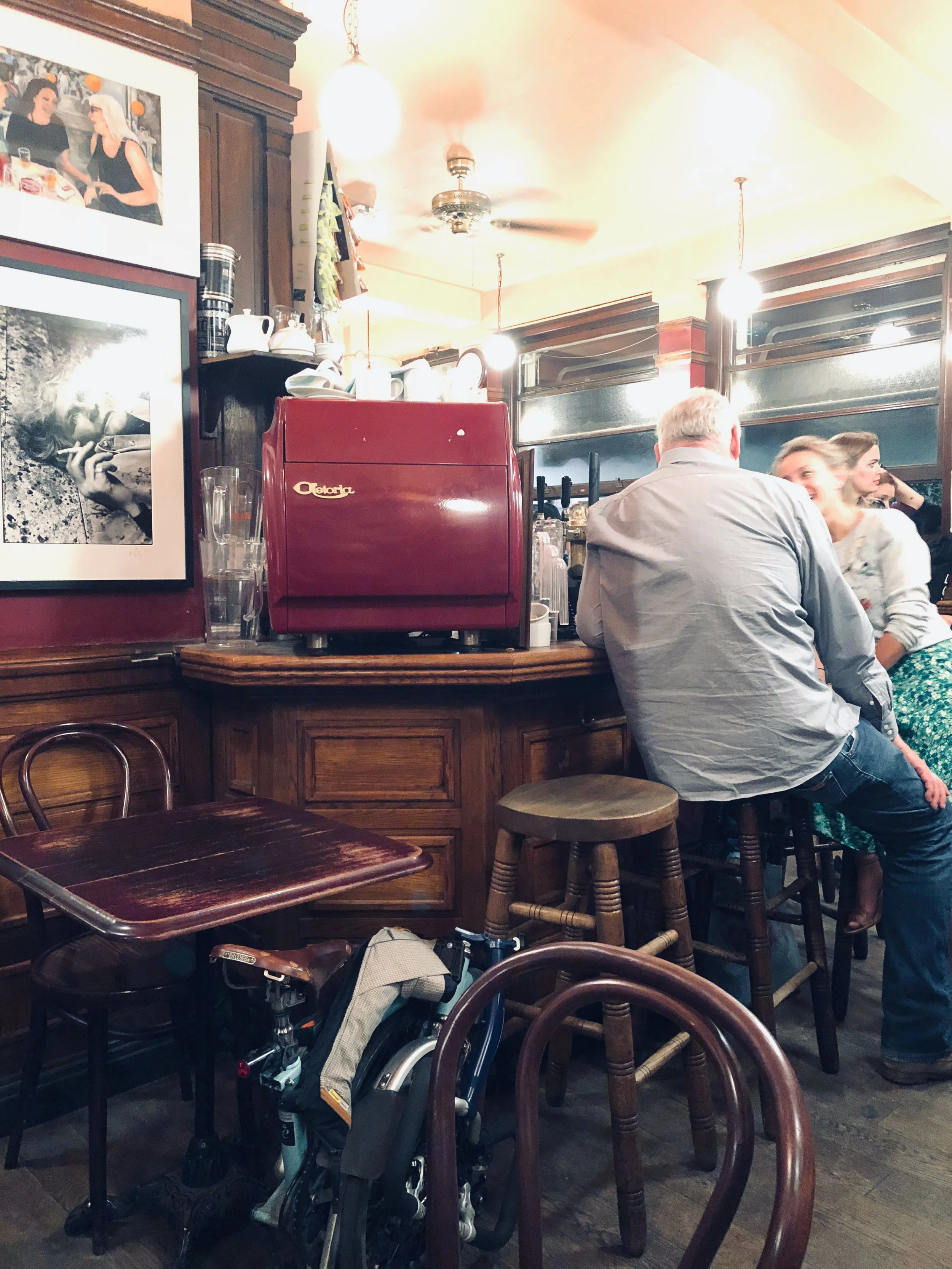 Interior of a cozy bar or restaurant with wooden furniture and warm lighting, featuring a red espresso machine, framed black-and-white photographs on the wall, and a group of people sitting at the bar, including a man with gray hair and a woman with 