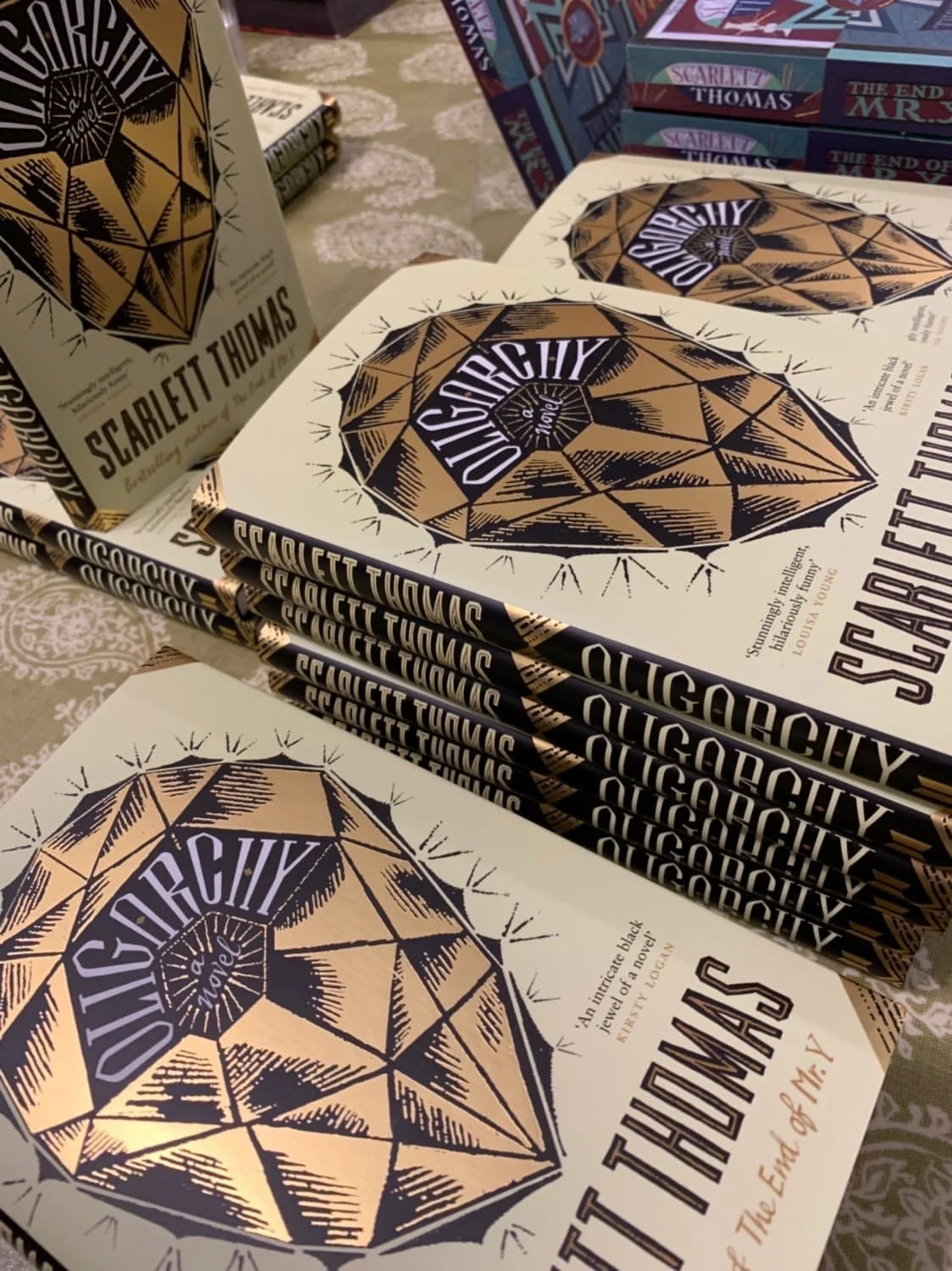 Stacked copies of the book Oligarchy by Scarlett Thomas with the cover featuring a stylized, hand-drawn diamond shape design in black and gold.