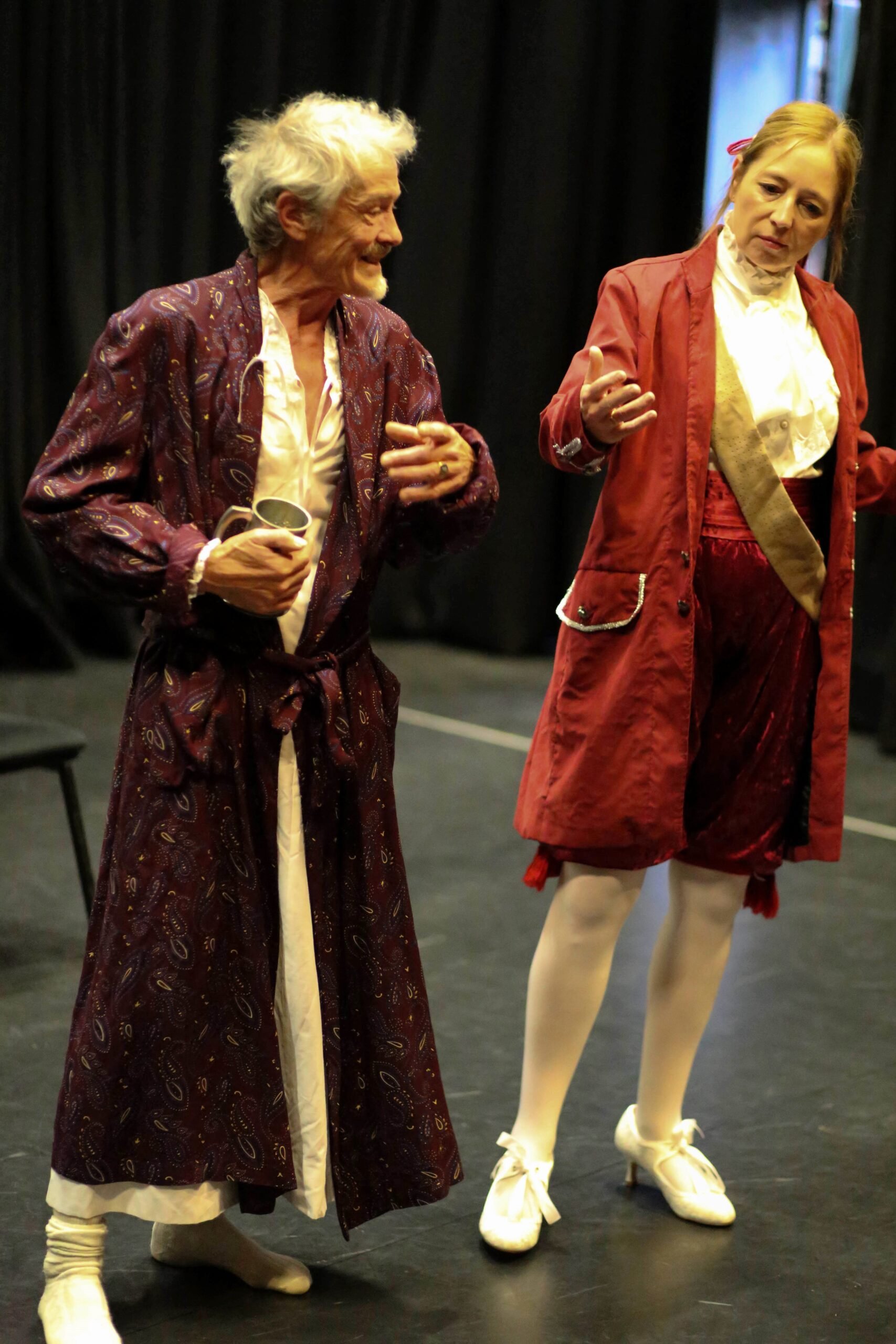 Two actors dressed in old-fashioned costumes stand on a stage with black curtains behind them. The elderly man on the left is wearing a patterned maroon coat and cream trousers, holding a cup, and smiling. The woman on the right has a serious express