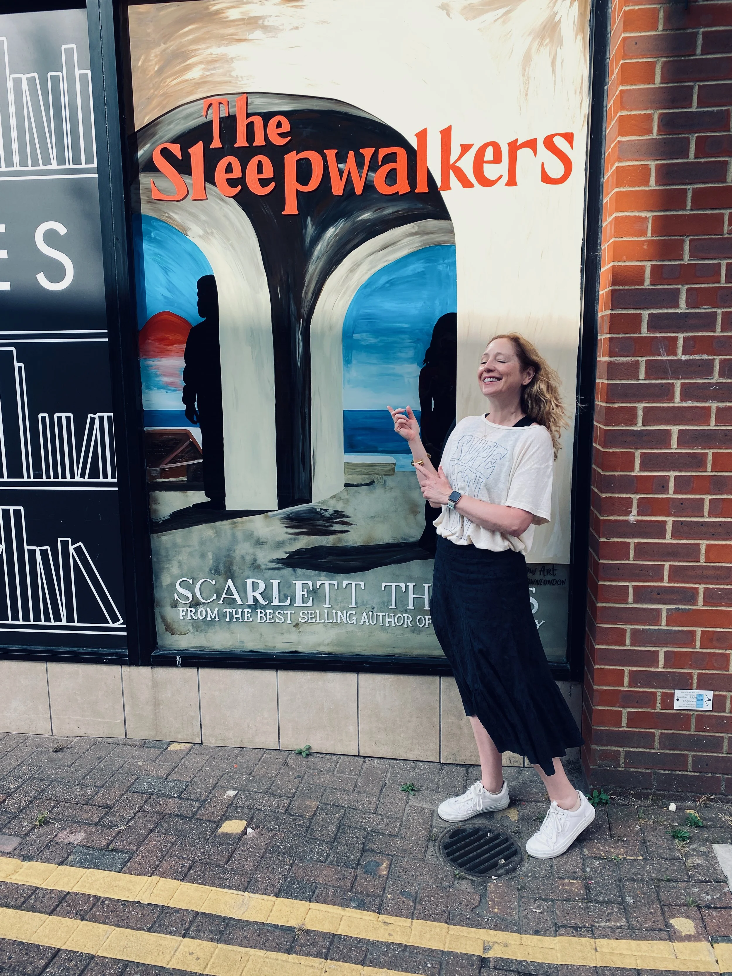 A woman in a white t-shirt, black skirt, and white sneakers smiling and pointing at a poster for the book 'The Sleepwalkers' by Scarlett Th.