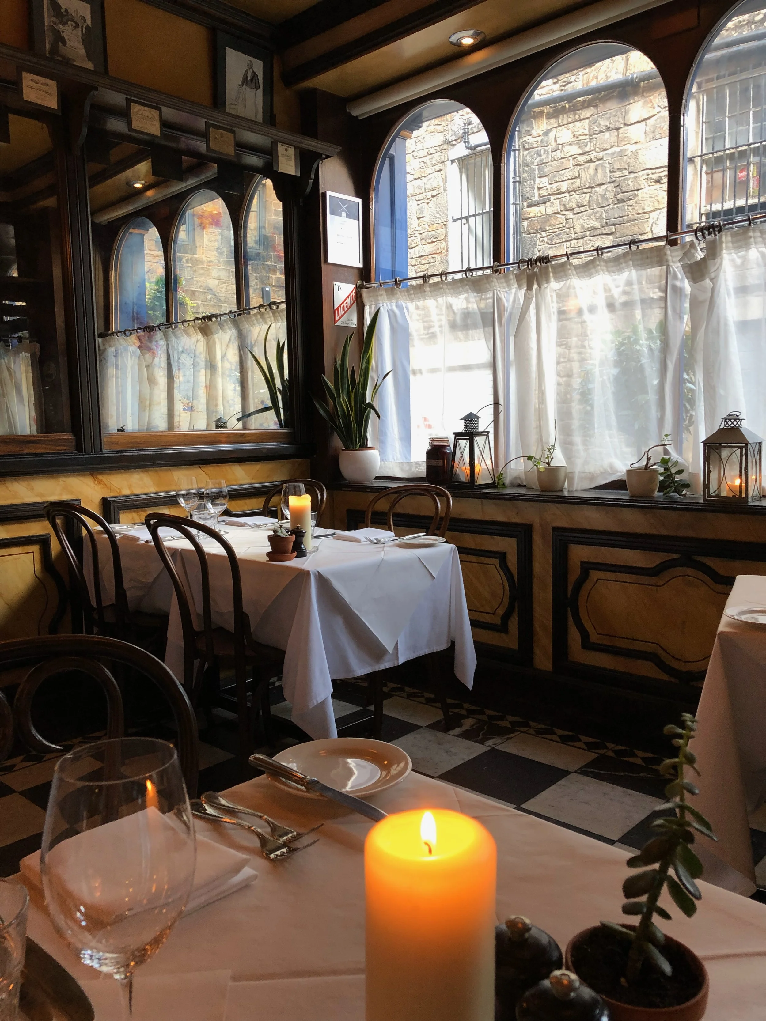Cozy restaurant interior with a table set for dining, lit by a candle and surrounded by potted plants, with large sunlit windows covered by white curtains.