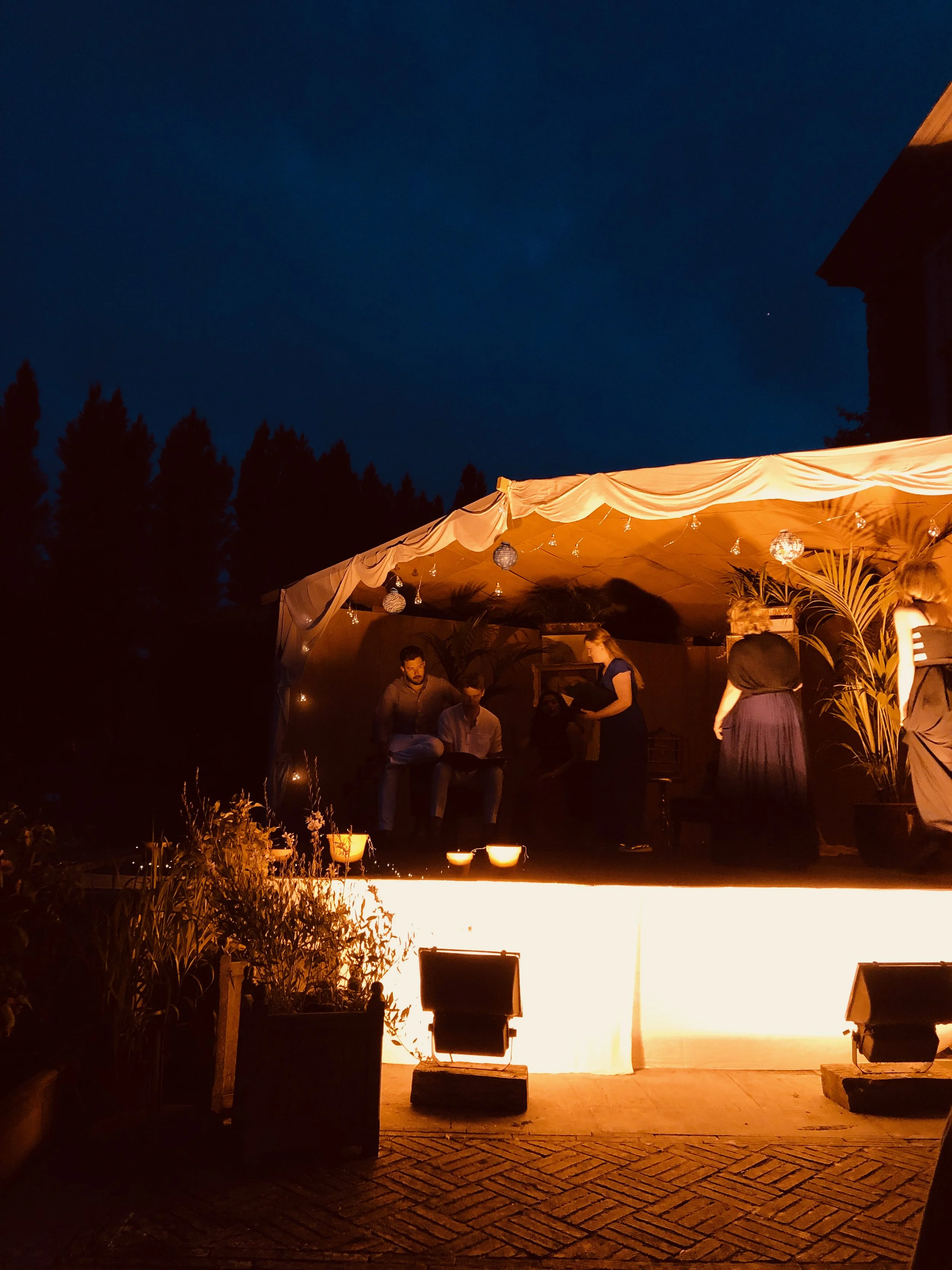 People performing on a small outdoor stage at night with string lights, plants, and a dark sky in the background.