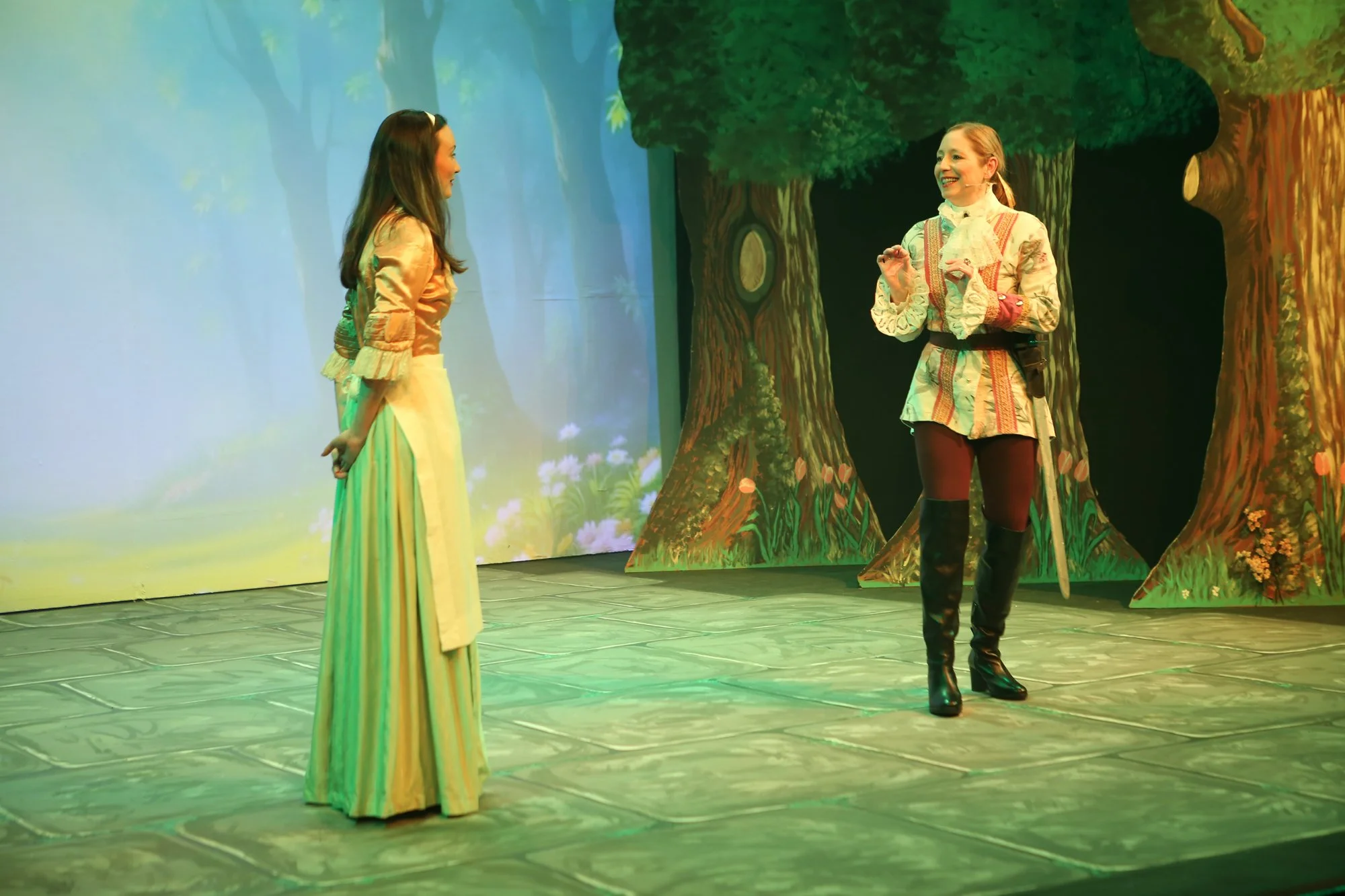 Two women in costumes on a stage with a forest backdrop. One woman is wearing a long cream-colored dress, and the other has a colorful tunic with tights and tall black boots.