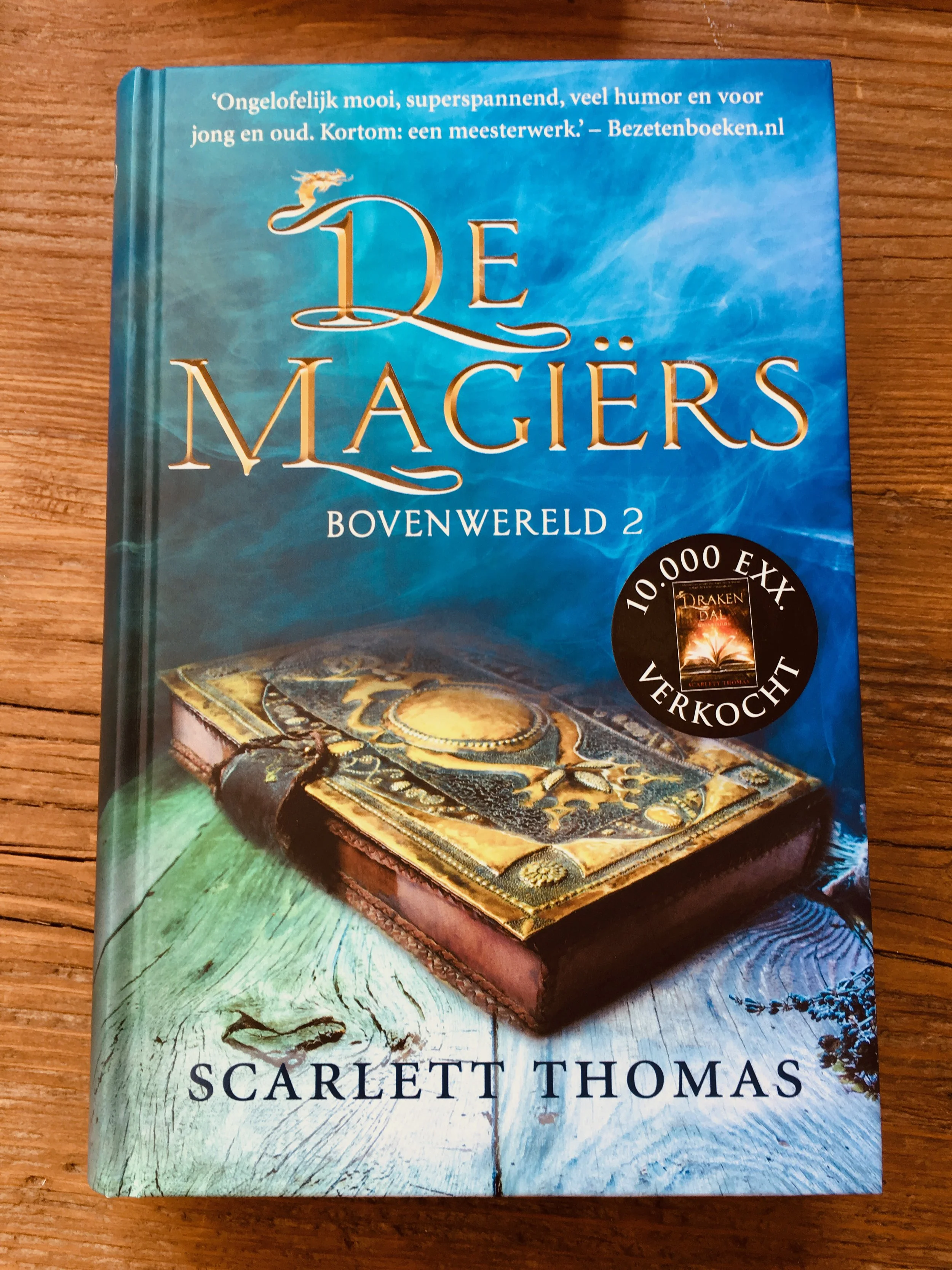 A book titled "De Magiërs" by Scarlett Thomas, with a blue sky and swirling clouds on the cover, featuring an ornate magic-themed box placed on a wooden surface.