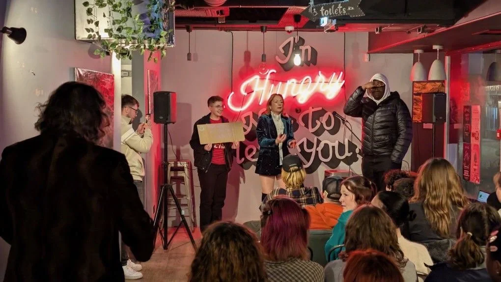 A group of people performing on stage in a venue with a red neon sign saying 'I'm Happy to See You!' in the background, and an audience watching.