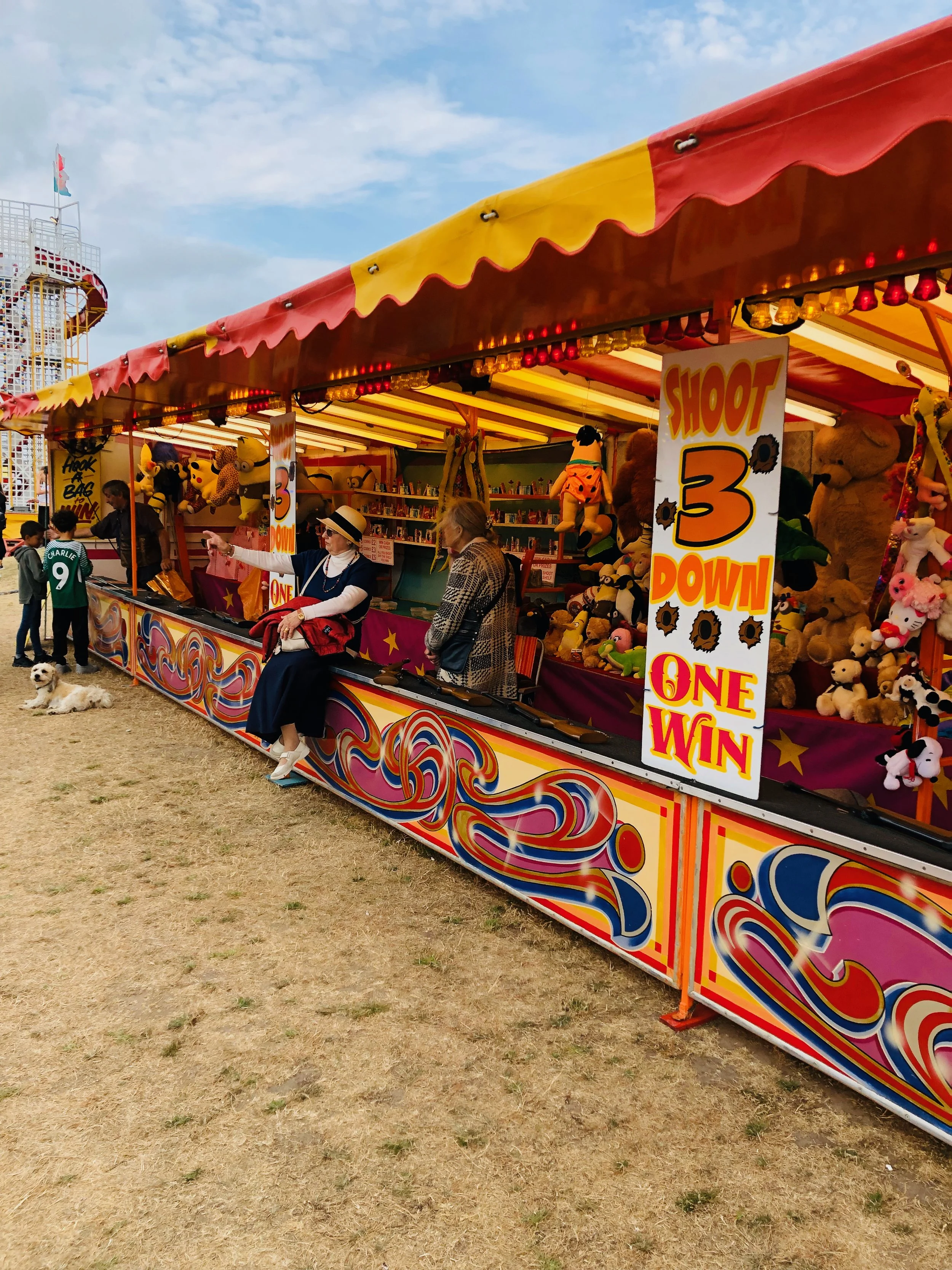 Colorful carnival game booth with plush toys, a sign that reads 'Shoot 3 Down, One Win,' and two women sitting on the counter, one wearing a large sunhat and sunglasses. A dog is lying on the ground nearby, and children are standing in front of the b