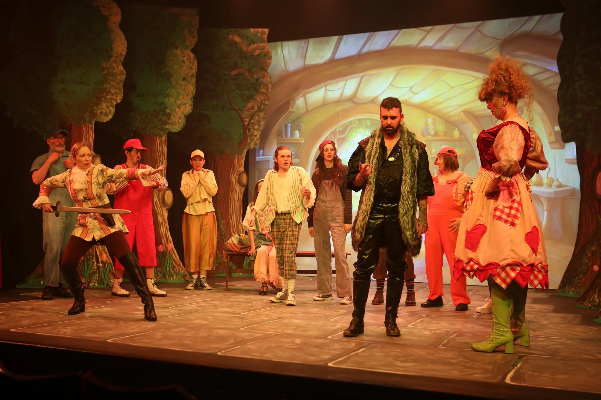 A theatrical stage with actors in colorful costumes, depicting a scene from a play with a forest backdrop and fantasy elements.