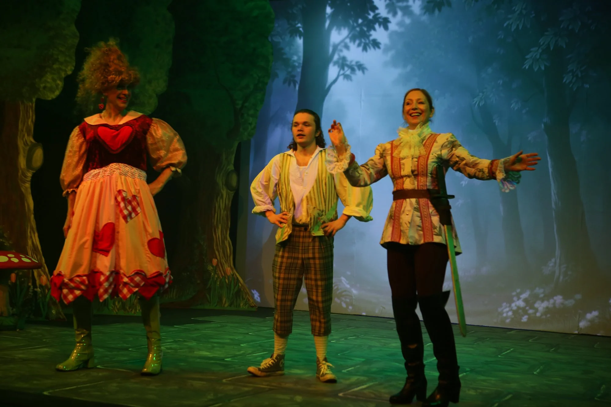 Three actors in colorful costumes performing on stage with a forest backdrop. The person on the left is dressed as a whimsical character with a vibrant dress and curly hair. The person in the middle wears a plaid outfit, and the person on the right i