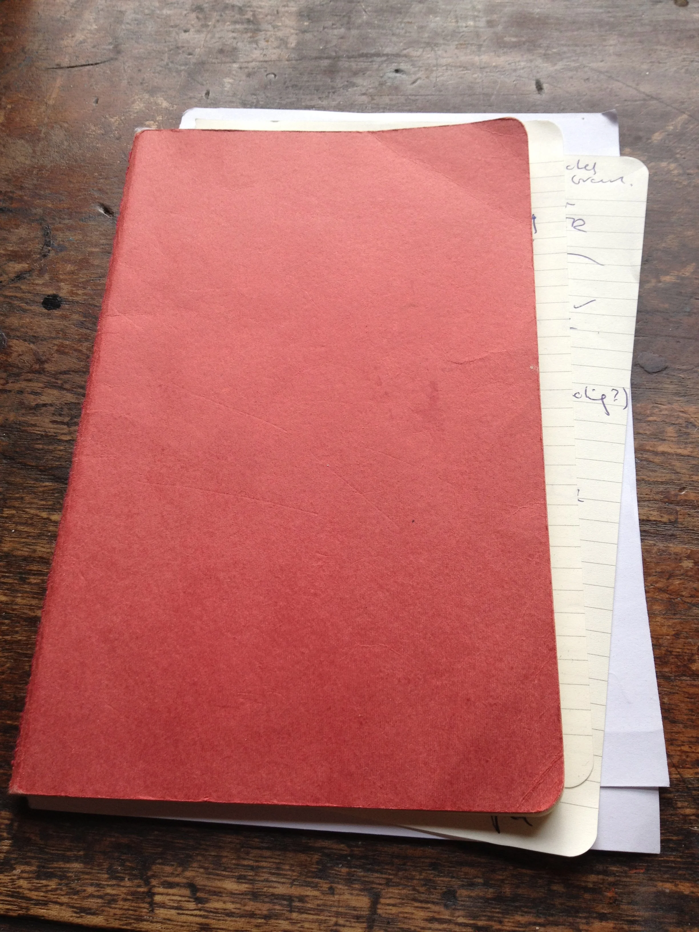 A red hardcover notebook placed on top of lined paper notes on a wooden table.